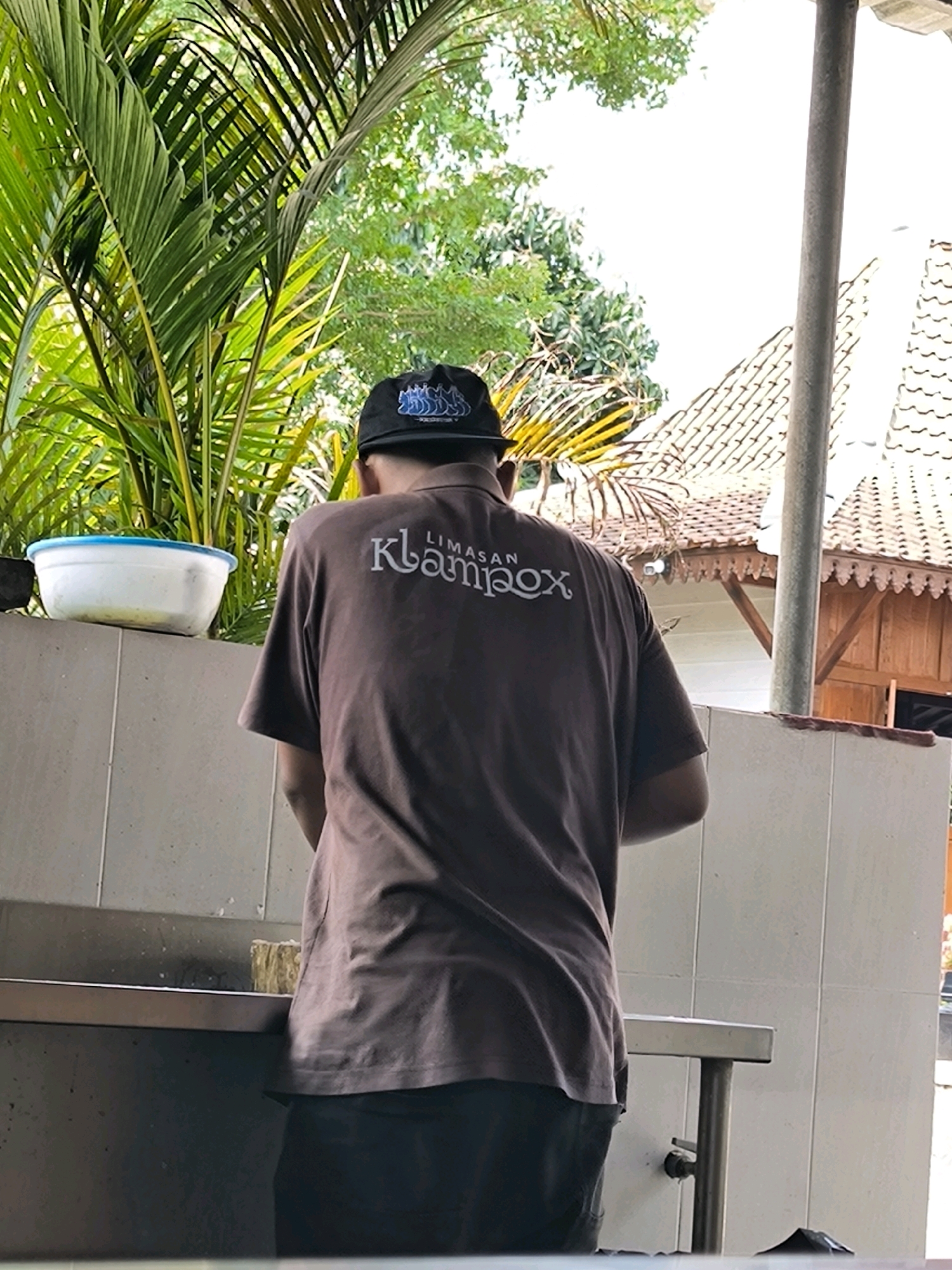 A person wearing a brown t-shirt with "LIMASAN klampox" printed on the back and a black baseball cap is standing outdoors, facing away from the camera. The person appears to be washing dishes or performing some task at a stainless steel counter. To the left, a white bowl with a blue rim sits on a light-colored wall. Behind the person, palm fronds and lush green foliage are visible, with a hint of a tiled roof in the background. The scene is lit by natural daylight.