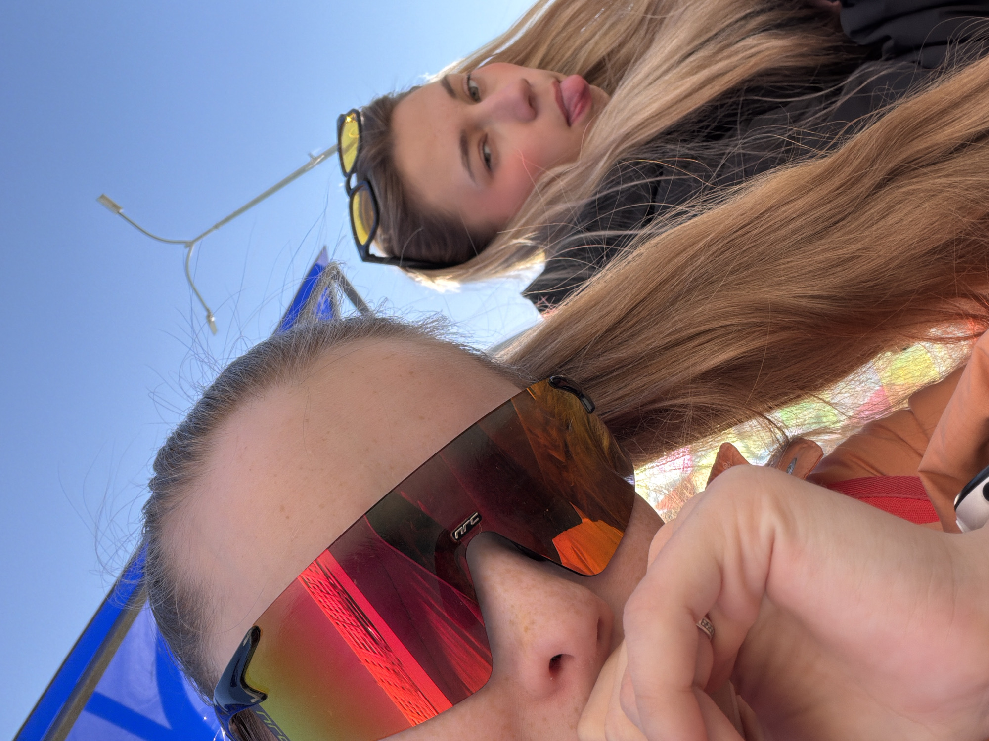 Two young women are depicted in a low-angle selfie against a bright blue sky. The woman in the foreground wears reflective sunglasses, while the woman in the background sticks out her tongue. Long, light-brown hair is draped across both individuals. The setting appears to be outdoors, possibly near a city, based on the presence of a streetlight. Given the prompt's mention of Berezniki, Russia, it can be inferred that the image was taken in or around that city.