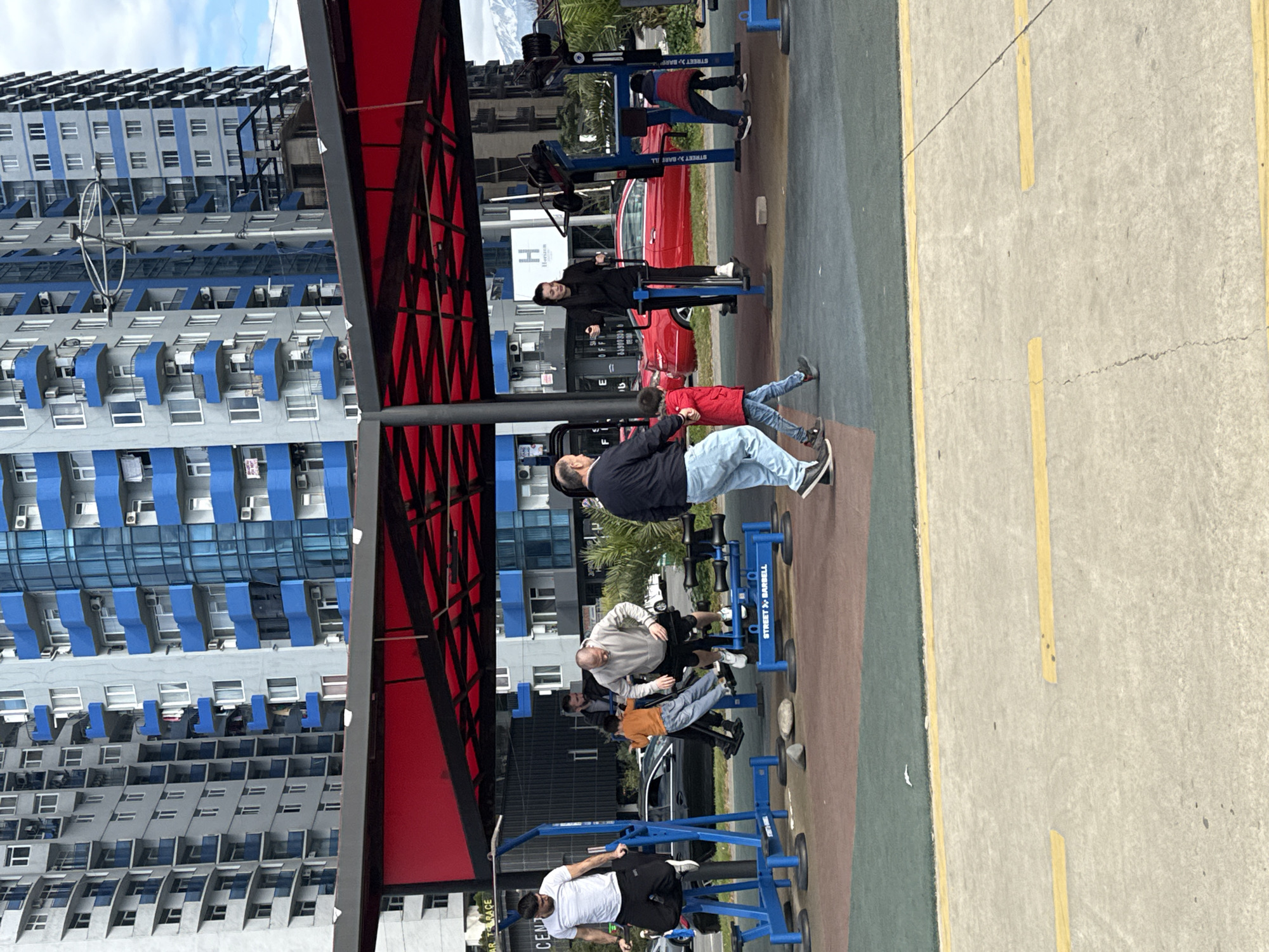 This image captures an outdoor public gym scene in Batumi, Georgia, on a bright, partly cloudy day. Several individuals are engaged in exercise activities.
In the foreground, a man in a white T-shirt and black pants is performing pull-ups or dips on a blue "STREET BARBELL" labelled machine. To his right, another man in a light grey hoodie and jeans is using a different blue gym apparatus. Behind them, a child in an orange top is sitting on an exercise machine, with an adult standing nearby. Further back, a man in a black jacket and light blue jeans is actively using a leg press-style machine, also part of the "STREET BARBELL" series.
The gym area is covered by a striking red canopy or roof structure. Behind the exercise equipment, a red car is parked, and a large "Horizon" sign with an "H" logo is visible on a building.
The background features towering, modern residential or commercial buildings characterized by their grey and blue facades, numerous balconies, and visible air conditioning units. A concrete pathway with yellow demarcation lines runs alongside the gym area. Additional visible text includes "AR TRACE" and "CENT" (partially seen on a sign) and variations of "STREET BARBELL" on the equipment.