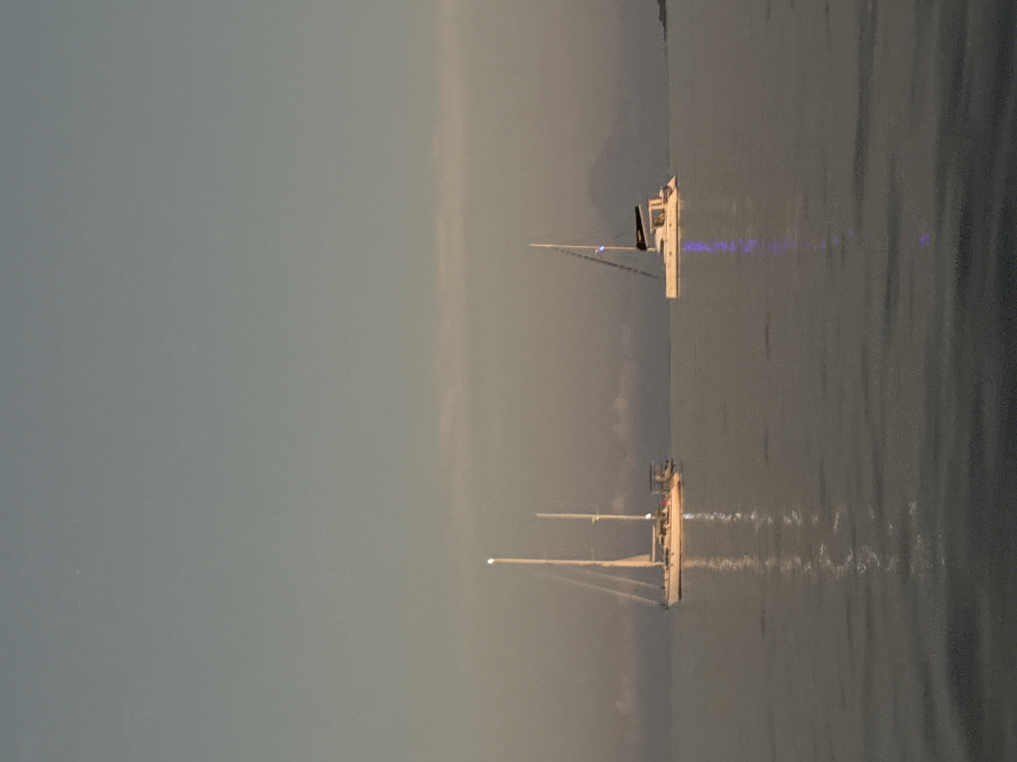 This is a medium-shot, vertical, still photograph of two sailboats on a calm body of water, likely the ocean, under a hazy, overcast sky. The lighting suggests either dawn or dusk. The sky is a gradient of pale blue and gray, with soft, indistinct clouds. The water is smooth and reflects the sky and the boats.
The sailboat on the left is a monohull with two masts, appearing to be a sailing yacht. It is white with some darker details on its deck. The masts are tall and slender, with rigging visible. A light is visible at the top of the taller mast. The boat is positioned in the left half of the frame.
The sailboat on the right is a catamaran, characterized by its two parallel hulls connected by a deck. It is also white with a dark cabin structure and a single mast. A blue light is visible on the side of this boat, and its reflection shimmers on the water below, creating a vertical streak of light. This boat is situated in the right half of the frame.
In the distance, a faint, dark silhouette of a mountain or large landmass is visible on the horizon, suggesting the presence of an island or mainland. The overall atmosphere is serene and peaceful. The location is identified as Unknown, Indonesia.
