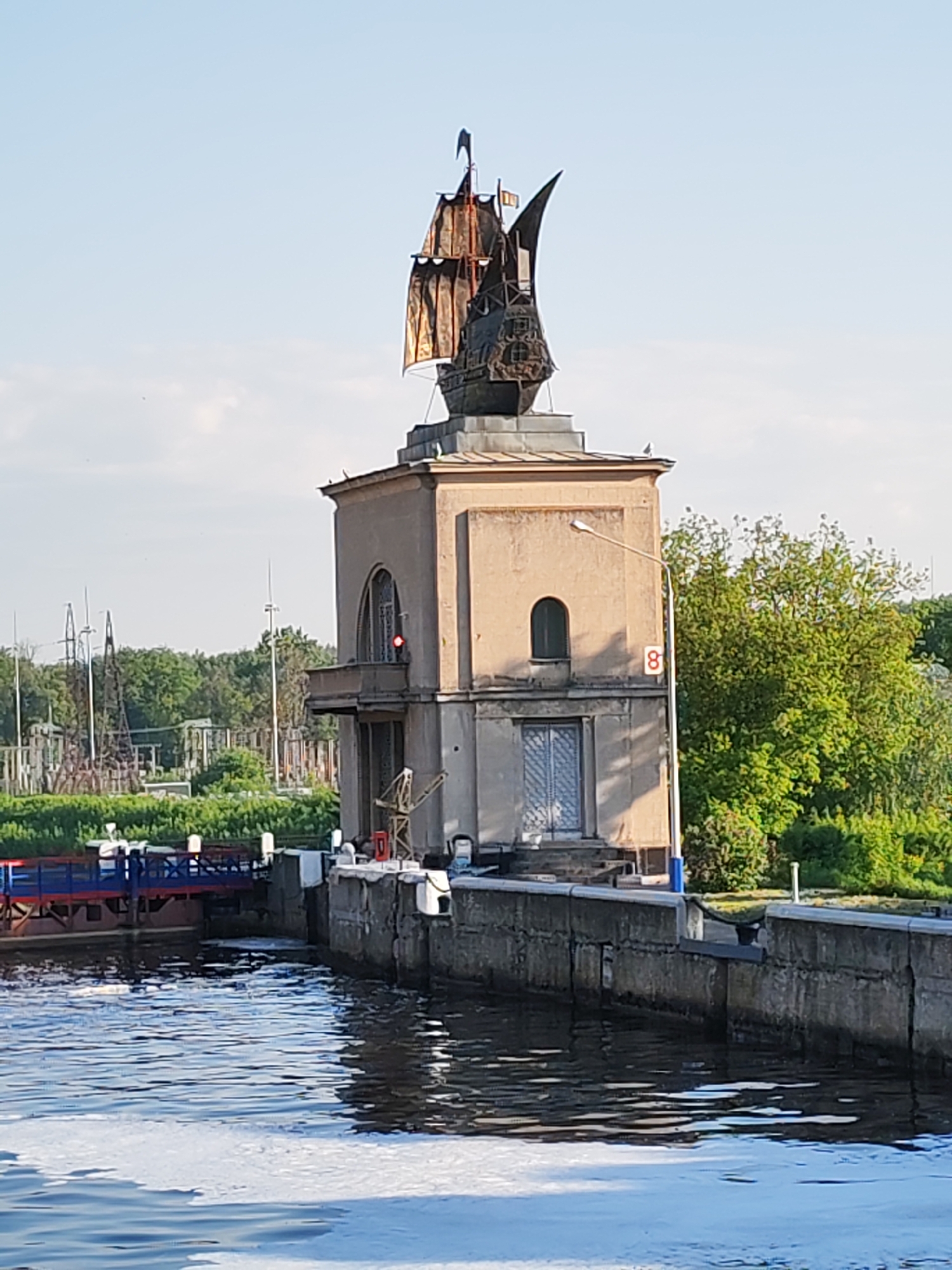 The image depicts a lock control tower at the Yakhroma Canal in the city of Yakhroma, Russia. The tower is a two-story beige structure with arched windows, a balcony, and a statue of a sailing ship on top. A red light is visible on the balcony. A lamppost with a speed limit sign (number 8) is located next to the tower. The lock wall is made of concrete blocks. A canal runs in the foreground of the scene, with a blue and red bridge-like structure on one side. Green trees and some industrial equipment are visible in the background.