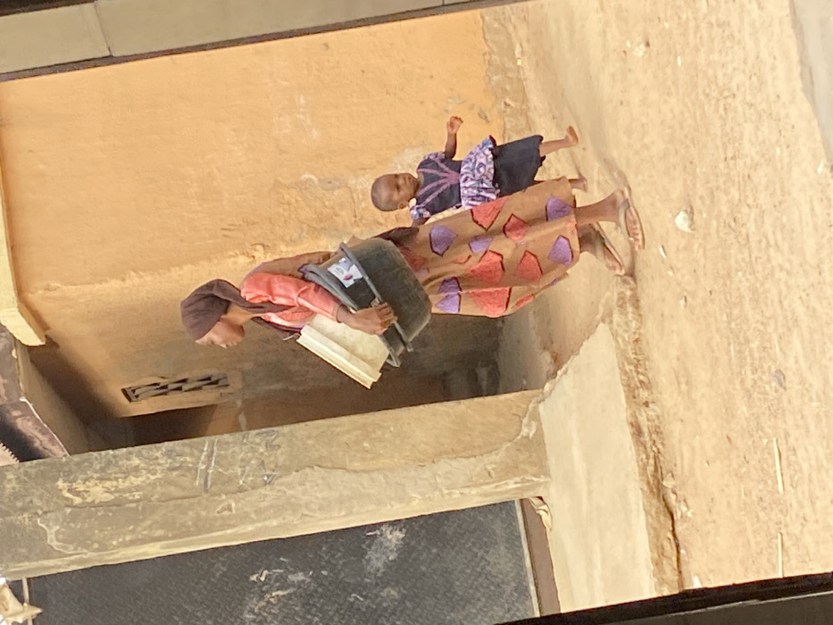 The image captures an outdoor scene in daylight, featuring two individuals: an adult and a young child. The perspective appears to be slightly elevated, looking down into a space bordered by architectural structures.
On the left side of the frame is an adult, likely a woman, facing towards the right. She is wearing a long dress or skirt with a brown or tan background patterned with purple and reddish geometric shapes. Her top is a lighter, possibly pinkish or orange, long-sleeved garment, and she has a dark brown head covering. In her left arm, she holds a dark-colored, possibly black, bucket or container. Tucked beneath the bucket, also held by her left hand, is a flat, rectangular object, which could be a book, a tray, or a board. Her feet are visible, wearing light-colored sandals.
To the right of the adult, slightly behind her, stands a young child. The child is wearing a dark-colored dress with patterns, possibly in light purple or blue, visible on the bodice and skirt. The child's arms are bent at the elbows, and their hands are raised slightly, suggesting a gesture or perhaps movement.
The background consists primarily of building walls and ground. The upper left and central background feature a smooth, light brown or tan wall. To the right, the wall appears rougher and lighter in color. The ground beneath the individuals is an uneven, light-colored surface with patches of darker material, possibly dirt or debris. In the lower left quadrant of the image, below the adult, there is a dark, rectangular opening with a horizontal gridded pattern, which could be a vent or a small window. The very bottom of the frame shows a dark, textured surface, possibly part of a lower roof or pathway from the vantage point of the photographer.
The bright lighting suggests it is daytime, and the overall environment, including the architecture and clothing, hints at a warm climate. The activity appears to be an everyday moment, with the adult carrying items and the child accompanying them, possibly walking or standing still. There is no visible text in the image.