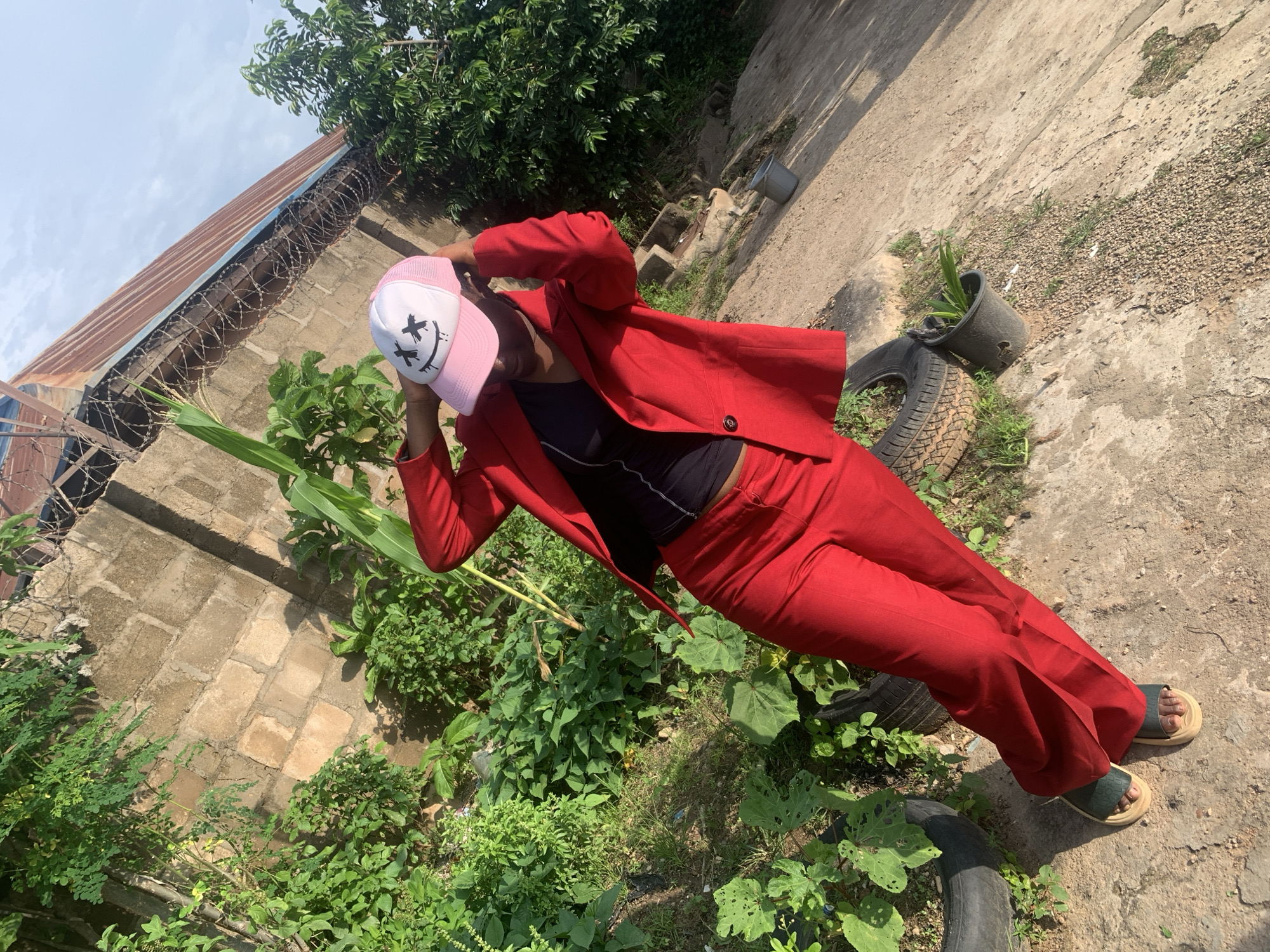 A young Black woman, wearing a red pantsuit and a pink baseball cap with a grimacing smiley face design, stands in an outdoor area. She is holding the brim of her cap with both hands and looking down. She is wearing a black top under the suit jacket and dark-colored slides on her feet. The background consists of a brick wall topped with barbed wire, lush green vegetation, and a corrugated metal roof. The ground is a mixture of concrete and dirt, with discarded tires and a black plastic bucket containing a plant. The scene appears to be in a residential area of Sabon Tasha, Nigeria, given the typical construction materials and vegetation seen. The lighting suggests it is daytime, with shadows cast by the woman and the surrounding structures.