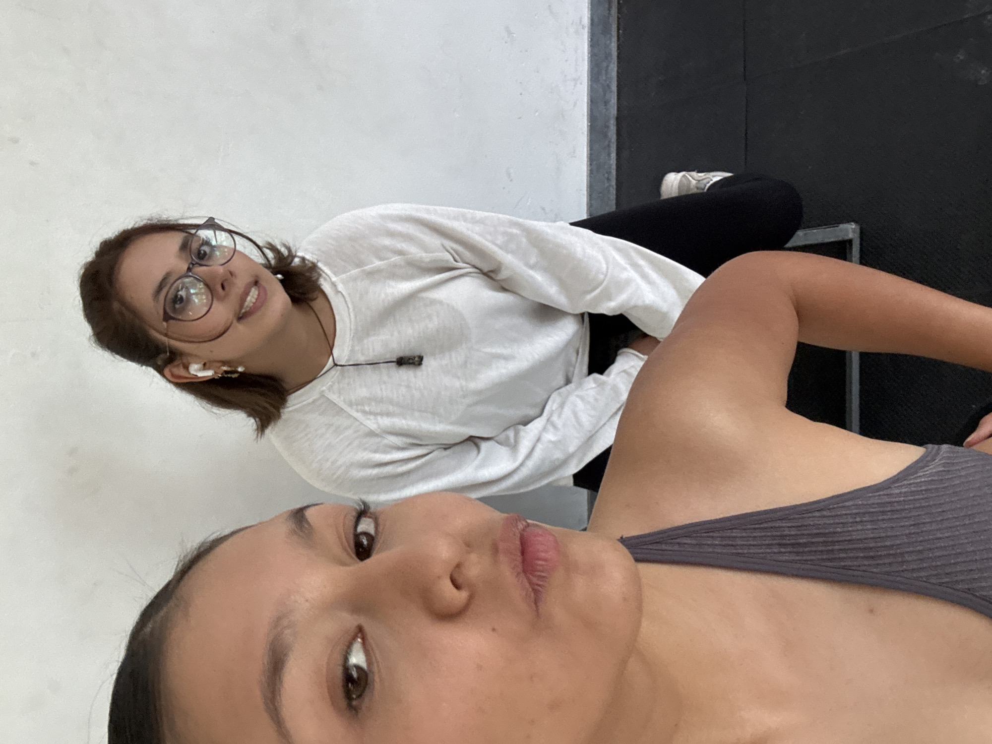 A tilted, close-up selfie captures two young women in an indoor setting, possibly a gym or fitness studio, characterized by a plain white wall and dark flooring or equipment. The image is taken from a low perspective.
In the immediate foreground, partially visible, is the woman taking the selfie. She has dark hair, brown eyes, and full, slightly parted lips. She wears a dark grey ribbed tank top or sports bra, with her shoulder and upper chest visible. Her expression is neutral.
Behind her, looking towards the camera with a relaxed smile, is a second woman. She has brown hair, wears dark-rimmed glasses, and has white wireless earbuds in her ears. She is dressed in a long-sleeved white t-shirt and black pants. A thin black cord is visible around her neck. Her feet, wearing white sneakers, rest on a dark surface, which appears to be a padded bench or mat. The background includes a dark vertical structure, possibly part of gym equipment or a wall trim.
The overall atmosphere is casual and relaxed. No text is visible in the image. The scene is located in San Andrés Cholula, Mexico.