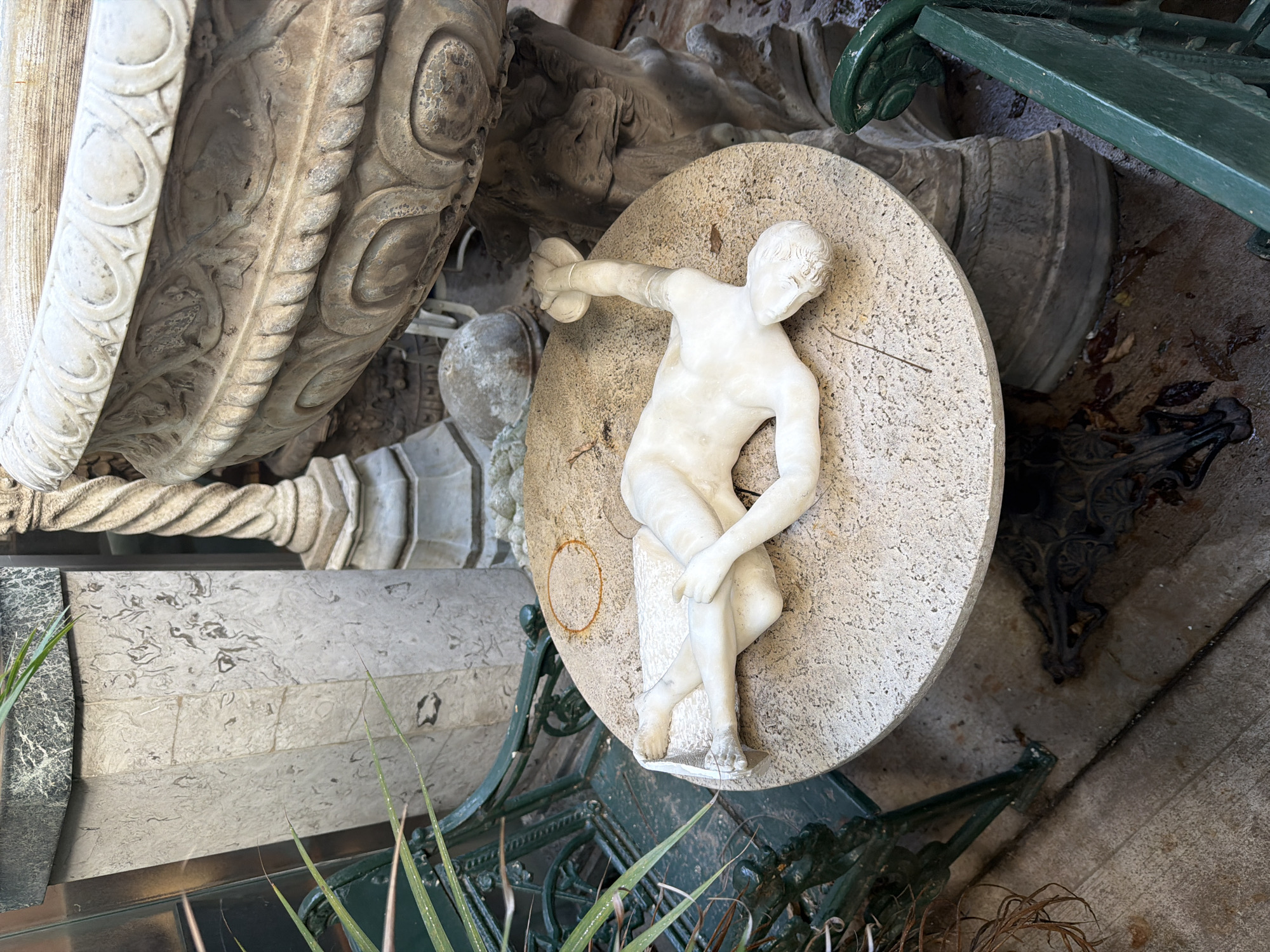 The image depicts a collection of architectural and sculptural elements, possibly in a garden or courtyard setting. The primary focus is a white marble statue of a nude male figure, poised in a dynamic stance, seemingly throwing a disc. The statue rests on a circular stone base with a rough, speckled surface.
Adjacent to the statue are fragments of heavily ornamented architectural details, including a section of a column with elaborate carvings and what appears to be part of a larger stone structure.
A green metal bench or structure is partially visible, adding to the cluttered and somewhat neglected atmosphere. The environment appears to be outdoors, with evidence of weathering and age on the stone and metal components.
Based on visual cues, this scene could be located in the city of Buenos Aires, Argentina, given the style of architecture and the prevalence of such historical elements in urban gardens or antique collections. However, without further context, precise location cannot be determined definitively.