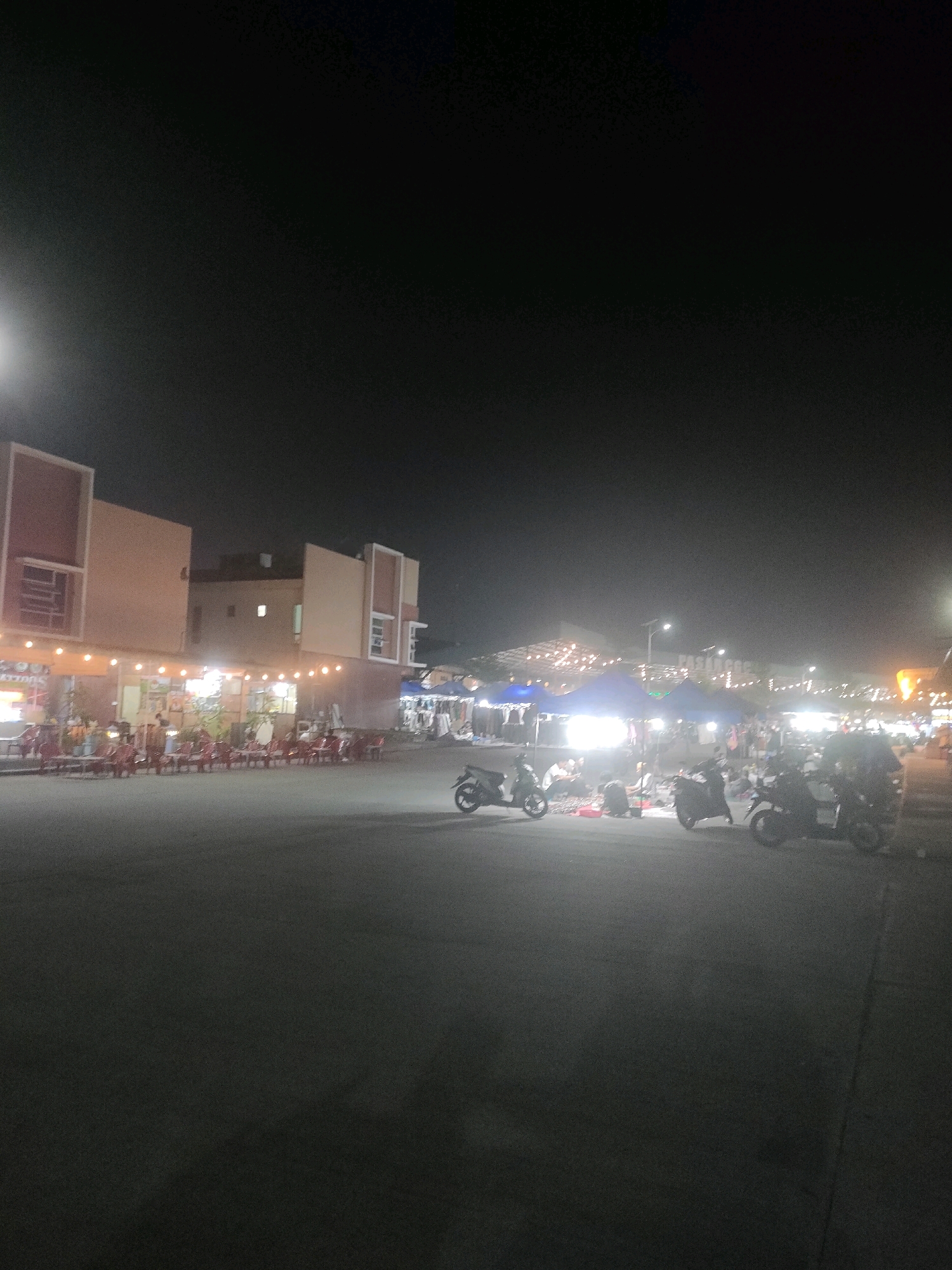 The image shows a night scene at a market in Batam, Indonesia. In the foreground, there is a paved area with several motorbikes parked. In the background, there are several market stalls set up under blue tents, illuminated by bright lights. People can be seen browsing and shopping at the stalls. The market appears to be busy and active. To the left of the market, there are buildings with storefronts that are also illuminated. The overall atmosphere is lively and vibrant, characteristic of a night market.