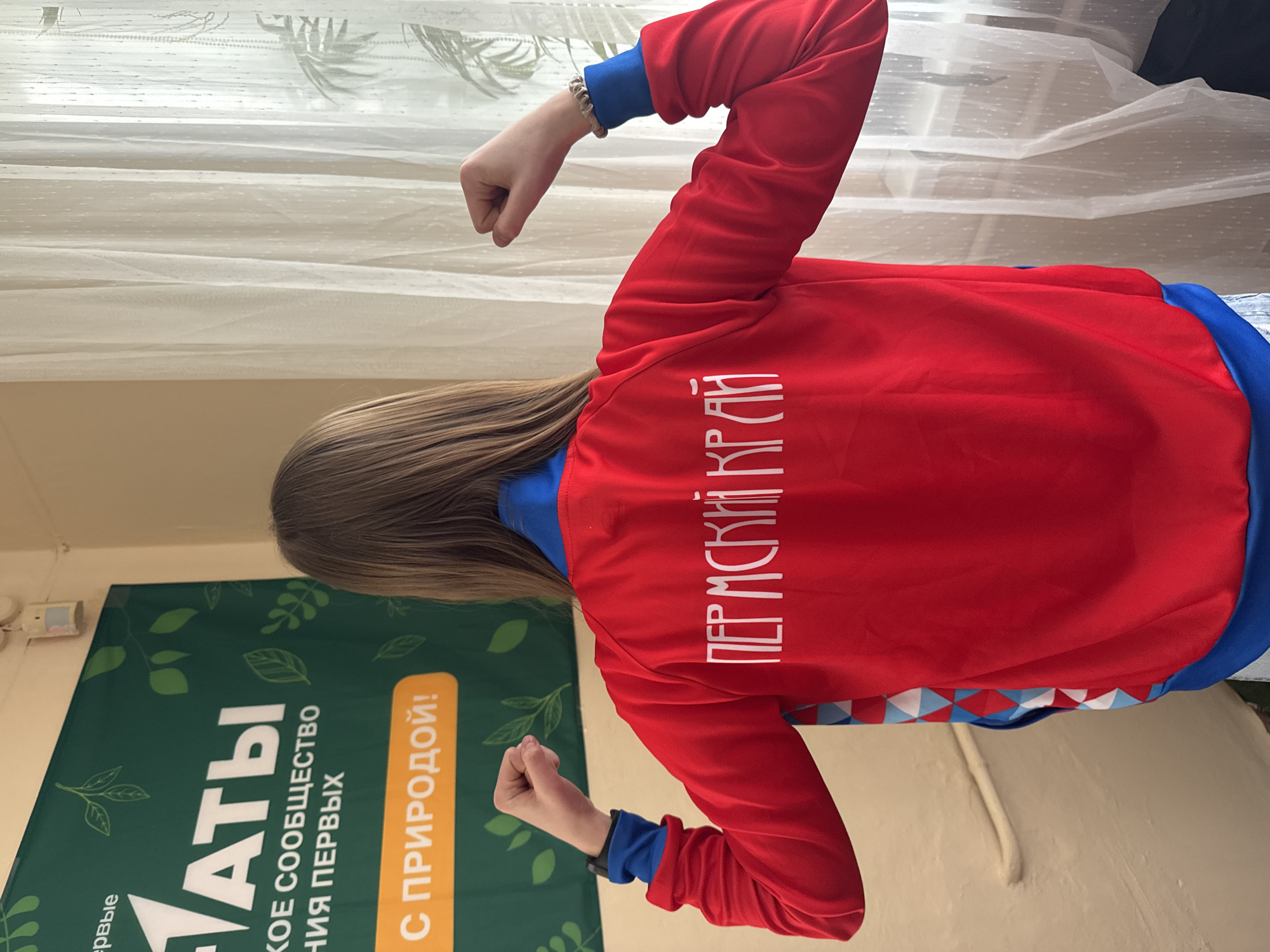 The image shows a person from the back, wearing a red and blue jacket. The back of the jacket has the words "ПЕРМСКИЙ КРАЙ" written in white. The person is standing with their fists clenched. Next to the person is a green banner with text written in Russian, including the phrase "С ПРИРОДОЙ!". A sheer white curtain hangs in the background. The location is likely Berezniki, Russia, as "ПЕРМСКИЙ КРАЙ" translates to "Perm Krai", a region of Russia, and Berezniki is a city within Perm Krai.