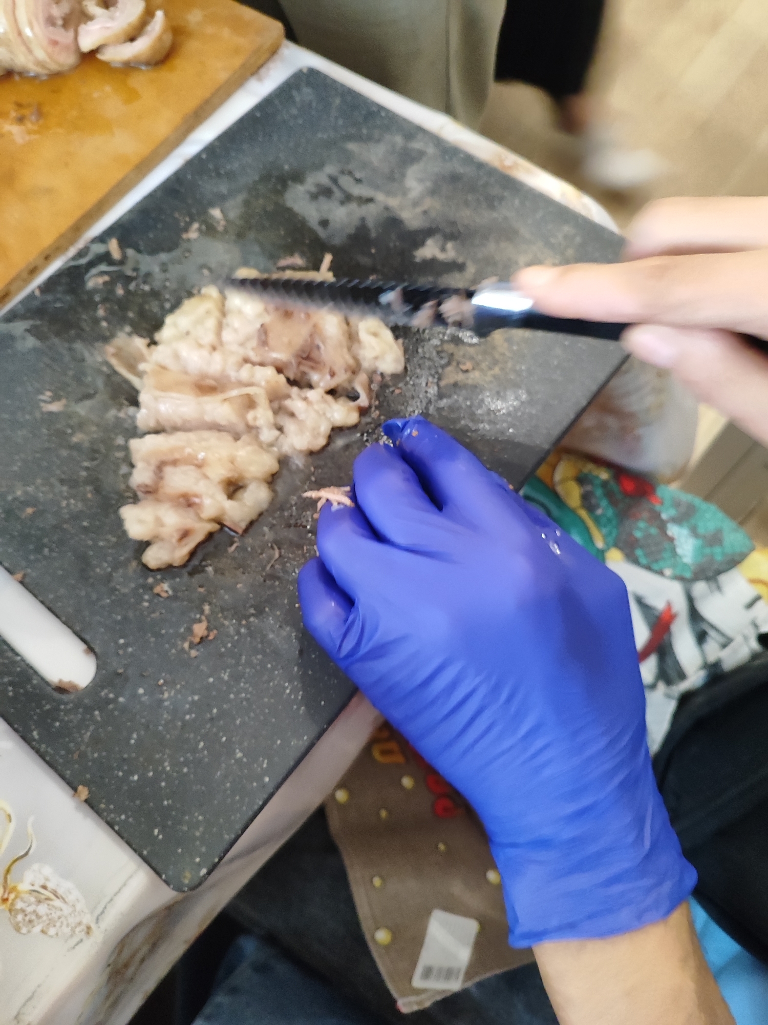 The image depicts a close-up view of a person wearing a blue glove and using a serrated knife to chop meat on a dark cutting board. Sliced meat pieces are visible on the board and on a wooden cutting board to the left. The environment appears to be a kitchen or food preparation area. There is no indication of the location within the city of Pavlodar, Kazakhstan.
