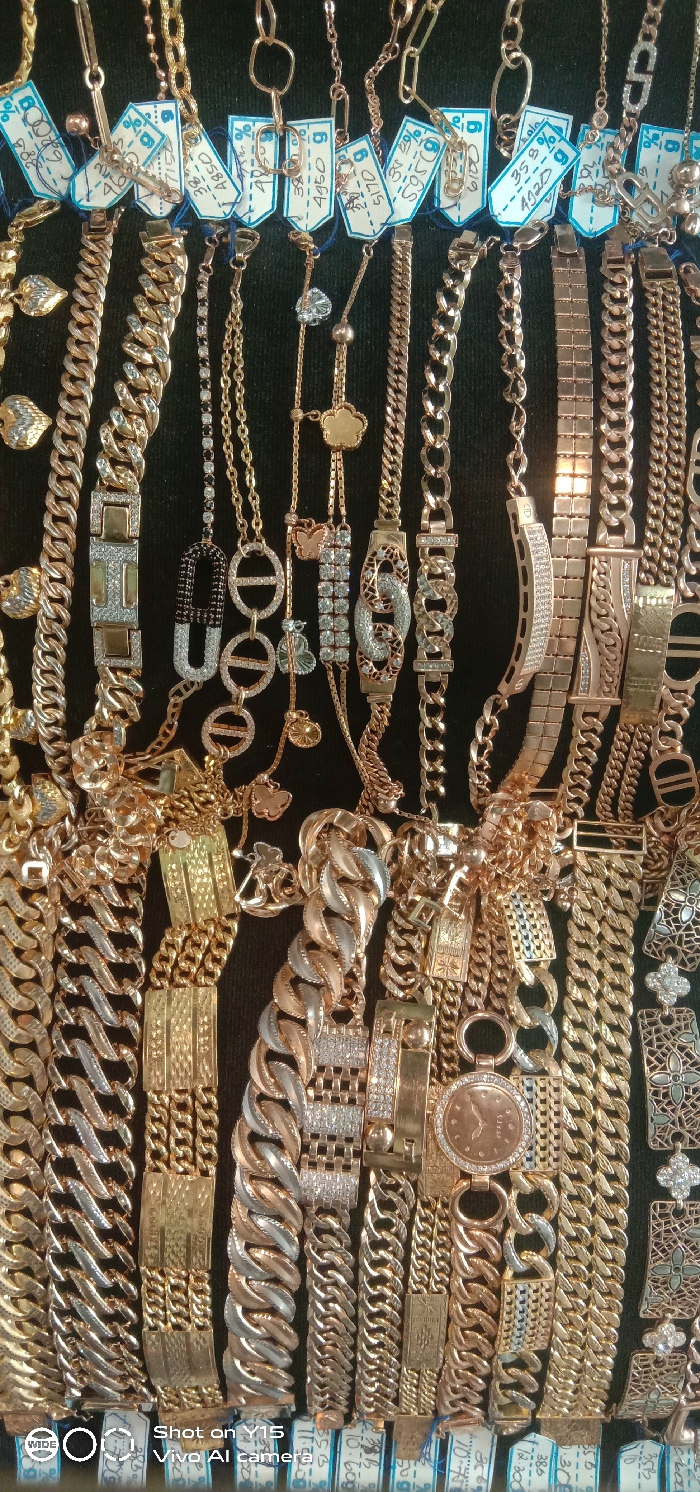 The image displays a dense collection of gold and rose gold-toned jewelry, primarily chains and bracelets, arranged on a dark, non-reflective surface, consistent with a retail display case in Maron, Indonesia. The jewelry pieces exhibit diverse designs, including curb links, woven patterns, geometric shapes, and various decorative elements like butterflies and stylized clovers. Several items feature embedded clear, faceted stones, likely diamonds or cubic zirconia, and some incorporate silver-colored metal accents. Small, white rectangular paper tags with blue handwritten text, including weights (e.g., 'g') and numeric values, are attached to multiple items, indicating inventory or pricing details. No individuals are present in the scene, and no active events are depicted.