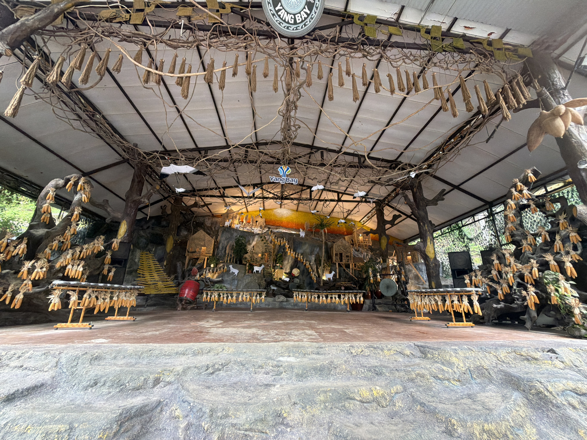 The image depicts a rustic outdoor stage area, likely a performance space or cultural exhibit. The stage is adorned with decorative elements made from natural materials, including dried corn husks and what appear to be bundles of straw or reeds hanging from the roof structure and branches. The background features a faux rock wall adorned with miniature structures, trees, and white goat figurines, suggesting a village or mountainous scene. Several racks display bunches of dried corn. On the left side of the stage, a large red drum is visible. The overall aesthetic is earthy and traditional, possibly representing a rural Vietnamese setting. The "Yang Bay" logo is visible at the top center of the frame, indicating the location is Yang Bay Park in Khanh Vinh, Vietnam. The scene appears to be empty of people.