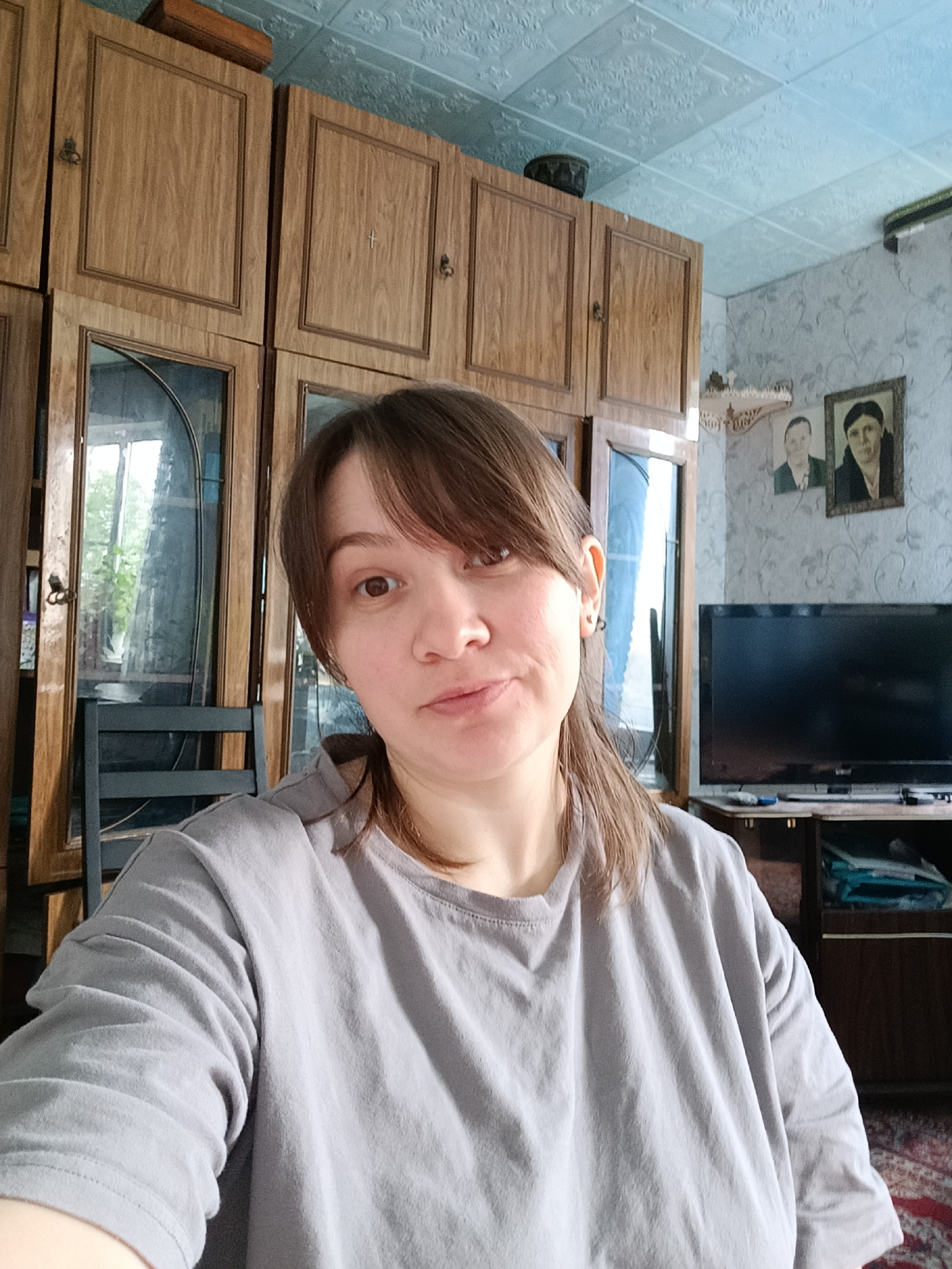 A young woman with brown hair and bangs is taking a selfie in what appears to be a living room. She is wearing a light grey t-shirt. Behind her, there is a large wooden cabinet with glass doors, a television on a stand, and two framed portraits hanging on the wall. The room has a textured ceiling and patterned wallpaper. The overall impression is of a cozy, lived-in space. The presence of framed portraits suggests a family home, and the style of the furniture and decor could indicate an older residence.