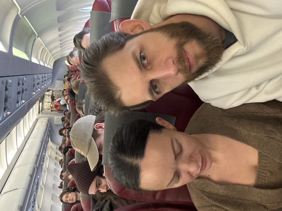 The scene depicts the interior of a commercial airplane cabin, located within the city of Novogorelovo, Russia. In the foreground, a male subject with a light brown beard and short brown hair, wearing a white hooded sweatshirt, looks directly at the camera with a neutral expression. Seated beside him to his left is a female subject with dark hair pulled back, wearing a brown knitted top and matching cardigan, looking downwards and slightly to her left. Both foreground subjects are seated in red-upholstered economy class seats.
The background shows a densely populated airplane cabin, with numerous passengers seated in rows of red and grey seats. Many individuals are visible, including one person wearing a brown beanie and another wearing a beige baseball cap. Further down the aisle, a crew member in a red uniform, identified as a flight attendant, stands near the rear galley or service area. The cabin features standard overhead compartments and interior lighting. No external view is visible through the windows.