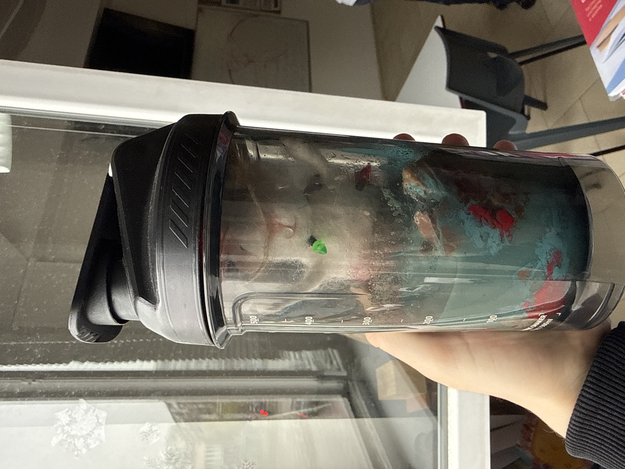A horizontal, eye-level shot depicts a person holding a protein shaker filled with liquid. The shaker is clear with black accents and volume markings. The liquid inside appears to be a mixture of teal, red, and other indeterminate colors. The person's hand and part of their arm, wearing a black ribbed sweater, are visible holding the shaker.
Behind the shaker, a window with a white frame is partially visible. The window shows a blurred view of what appears to be the outside. A radiator is below the window. In the background, elements of an interior space are visible, including a white surface, part of a black chair, and a white cabinet.
There is no indication in the image that specifies a location in Sofia, Bulgaria.