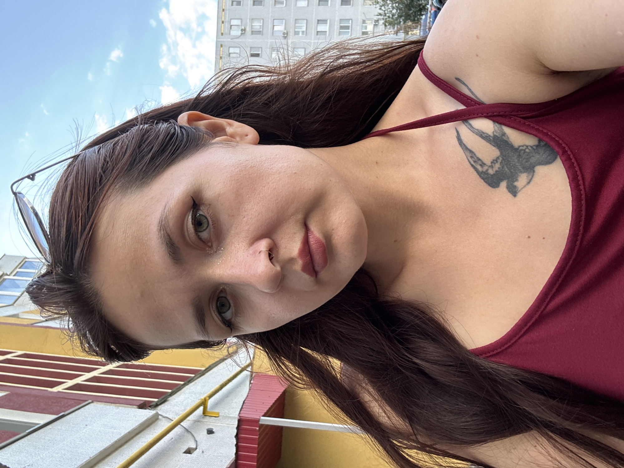 A medium shot selfie taken outdoors shows a woman with long, dark brown hair and green eyes. She is wearing a maroon tank top and has a bird tattoo on her chest. She is wearing a nose ring and has eye makeup on. Sunglasses are perched on top of her head. The background shows a building with red and yellow sections, and a tall, gray apartment building with many windows. The sky is visible, with some clouds. The location is the city of Naberezhnye Chelny, Russia.