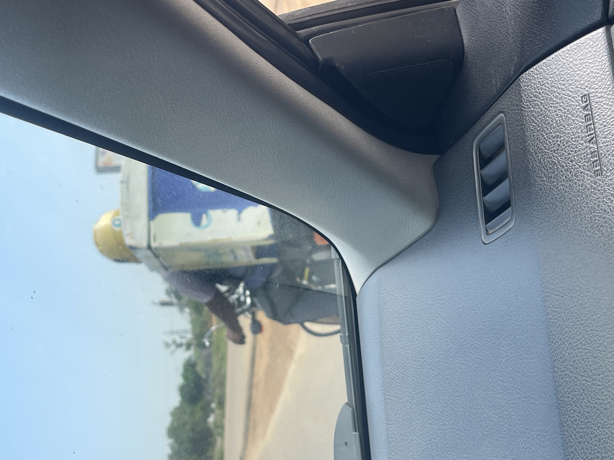 A person on a bicycle with a large cooler box on the back is visible from inside a car. The person is wearing a dark shirt and grey pants, and a yellow helmet. The cooler box is white with blue accents and appears to have a logo or branding on it. The bicycle is dark colored. The scene is outdoors, with a paved road and some vegetation visible in the background. The sky is clear and blue. The interior of the car, including the dashboard and a vent, is in the foreground. The image appears to have been taken in Kuchingoro, Nigeria.