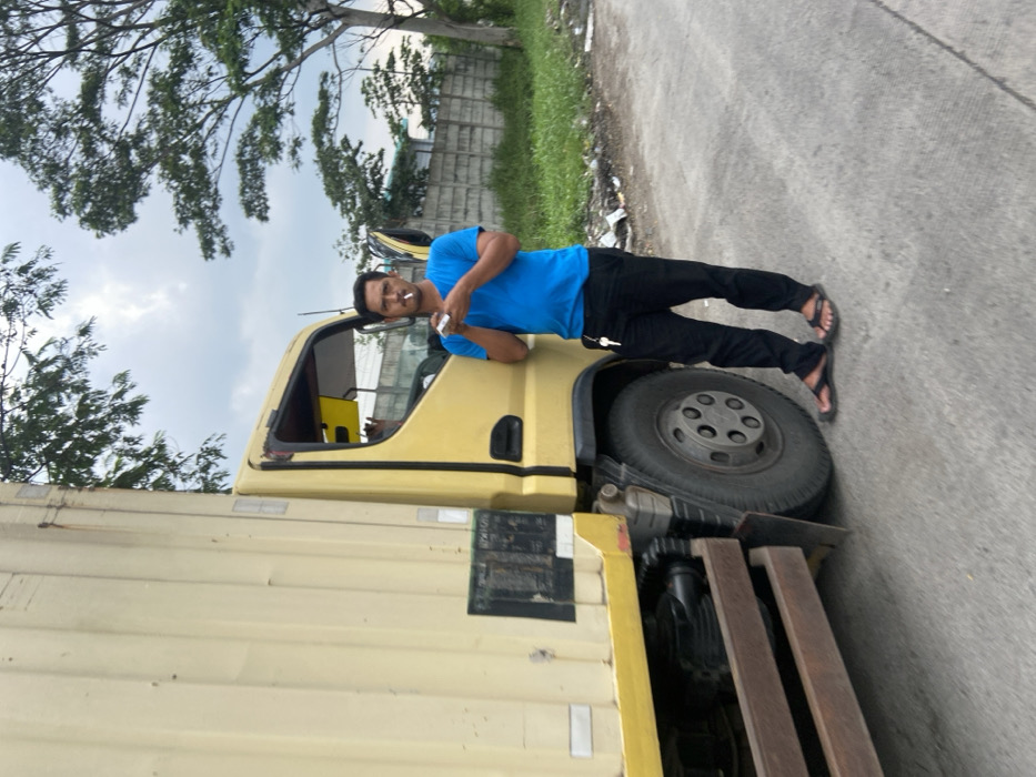 A man in a blue shirt and black pants stands next to a yellow truck. He is holding a cigarette in his right hand and appears to be smoking it. The truck is parked on the side of a paved road. The road is surrounded by vegetation. The man is looking directly at the camera. The location is Margaasih, Indonesia, based on the provided coordinates (latitude: -6.969, longitude: 107.549). The image appears to have been taken during the day, as the sky is clear and the sun is shining. The truck's cab door is open and a hand can be seen inside, possibly reaching for something. There are keys attached to the truck's side. The road is made of concrete, and it is dusty. The pavement is slightly cracked and uneven. There is a small amount of litter on the side of the road, near the truck. There is a concrete wall and a row of trees in the background.