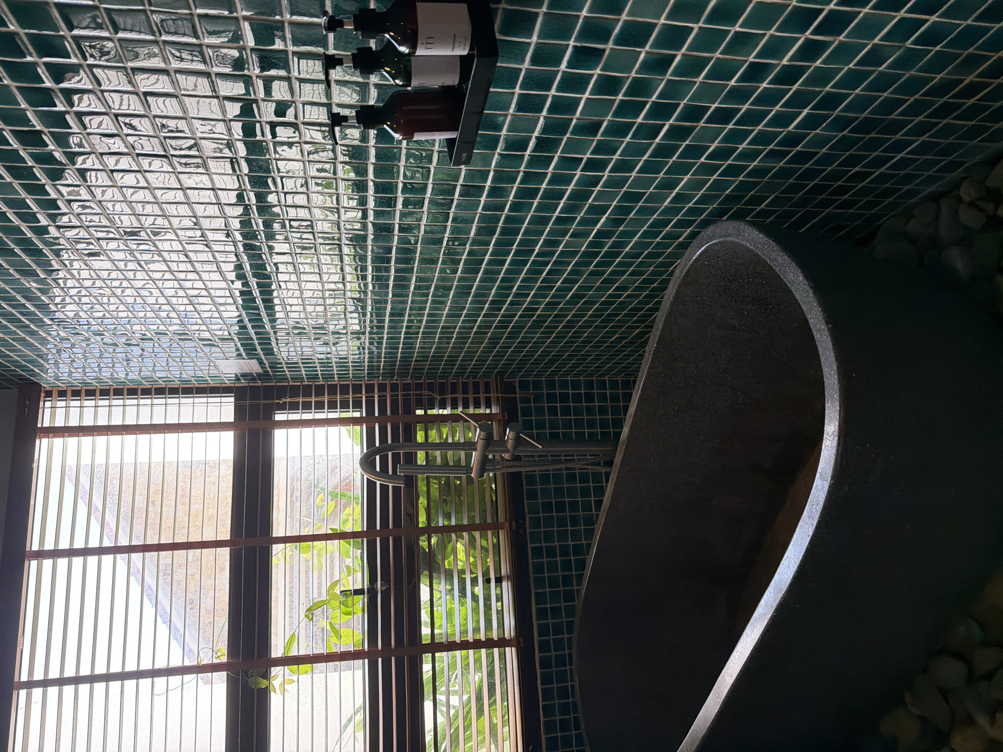 The image depicts a modern bathroom featuring a deep, dark gray bathtub situated next to a window covered by horizontal wooden blinds. The bathtub appears to be made of a textured material, possibly stone or a similar composite. Green, square mosaic tiles cover the wall behind the bathtub, creating a vibrant and reflective surface. On a small black shelf mounted on the tiled wall are three dark-colored pump bottles, likely containing toiletries. Outside the window, lush green foliage is visible, suggesting a connection to nature or an outdoor garden. The overall aesthetic is one of contemporary luxury with a touch of natural elements. The scene is set in a private bathroom within South Quy Nhon Ward, Vietnam.