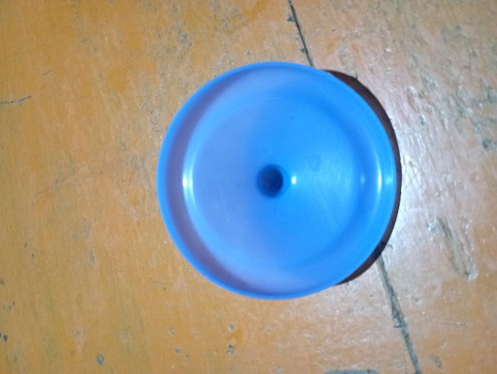 The image displays a single, circular, blue plastic object resting on a yellowish-brown, textured surface. The object is viewed from directly above, showcasing its slightly concave shape with a raised outer rim and a distinct, smaller circular hole in its center. The blue material appears to be semi-translucent or glossy, with subtle reflections visible on its surface.

The background surface shows signs of wear, including various scuffs, scratches, and darker patches, suggesting it might be a well-used wooden floor or a similar robust, earthy-toned material. A shadow is cast by the blue object onto the surface, primarily visible along its right edge, indicating an overhead or slightly off-center light source. The lighting appears natural, possibly indoor ambient light. There are no people, animals, or other prominent objects in the scene, and no specific activity is taking place. No text is visible in the image. The scene is static and focuses solely on the object and its immediate surroundings.