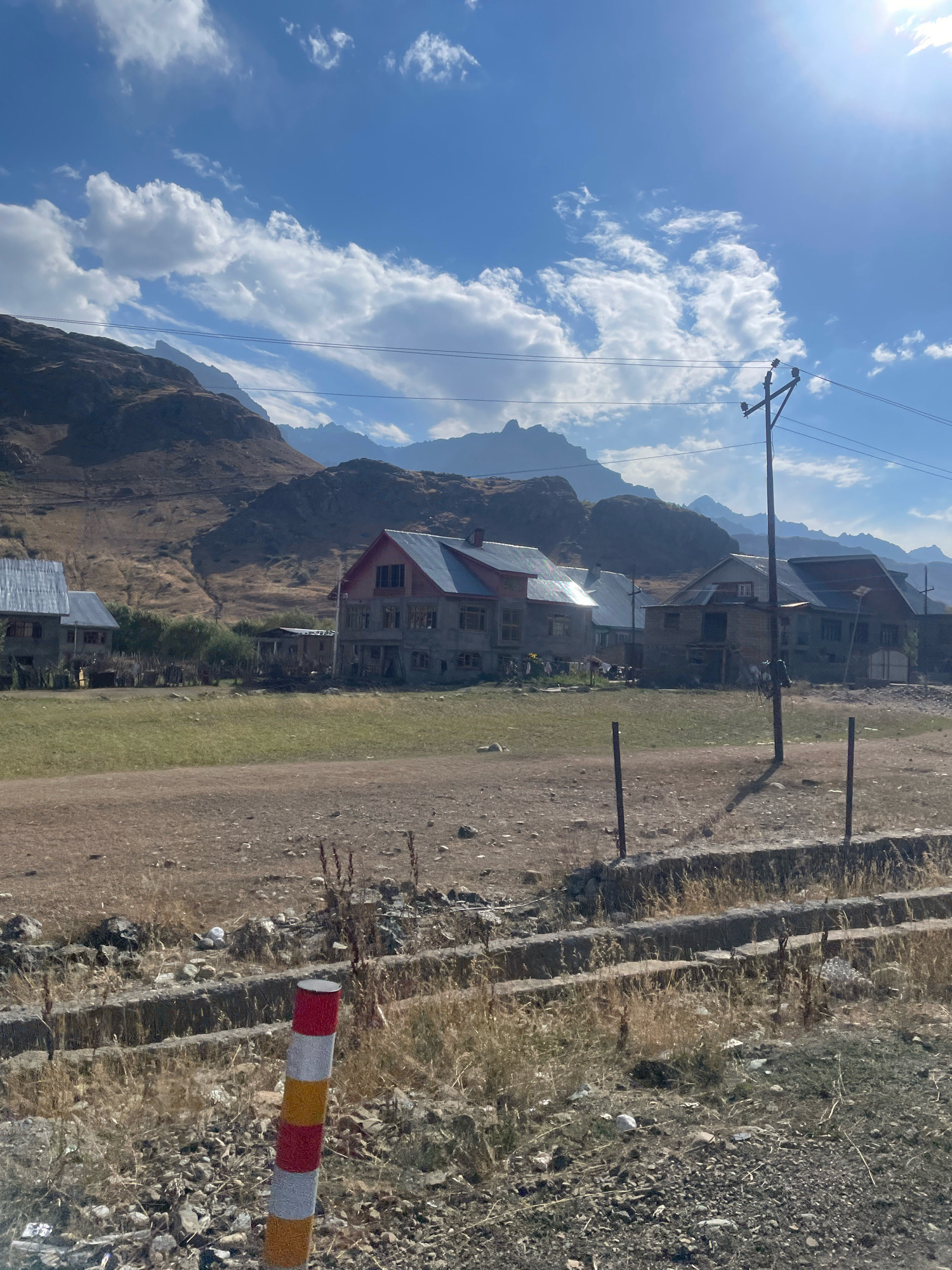 The image depicts a rural scene in the city of Dras, India, on a clear day. In the foreground, a red, white, and yellow striped pole stands vertically on a rocky, barren ground. The middle ground features a dry, grassy field and a shallow, dry ditch. Beyond the field, several multi-story houses are visible, some appearing to be under construction. The houses have red and grey roofs and concrete exteriors. In the background, rugged, brown mountains rise sharply against a bright blue sky with scattered white clouds. A utility pole with power lines crosses the sky near the mountains. The overall environment suggests a remote, mountainous region.