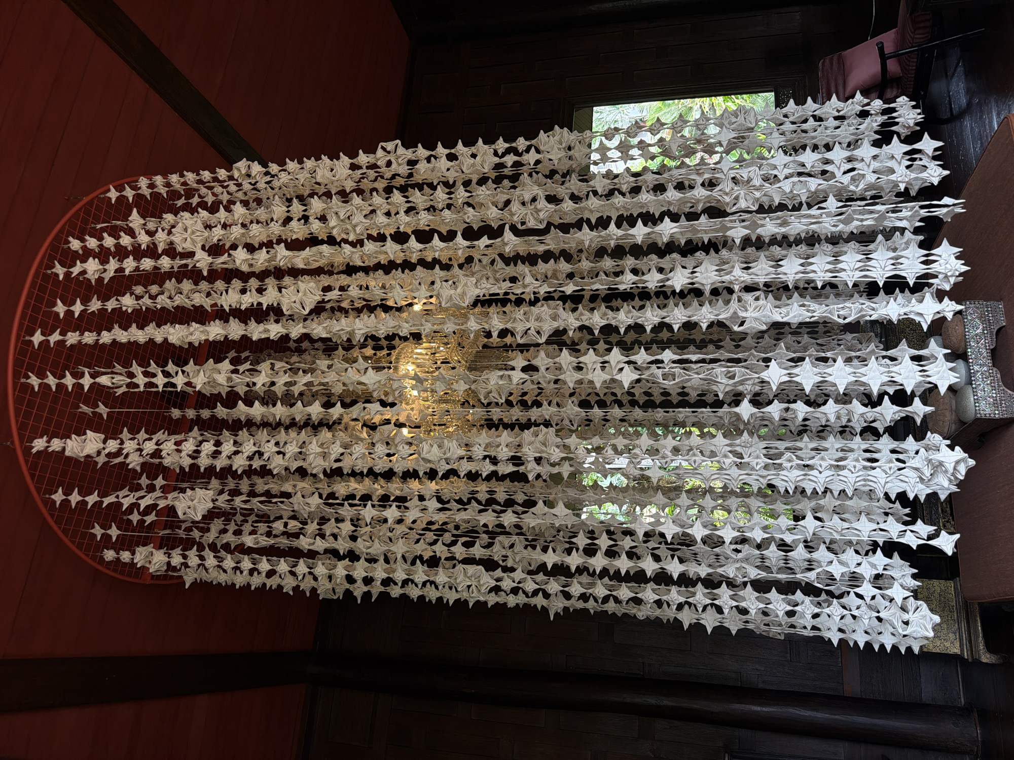 The image captures an interior shot of a chandelier-like art installation made of white, star-shaped paper or fabric elements suspended from a red circular frame against a dark wood-paneled wall and a red ceiling. The installation is a hanging curtain of intricately folded elements. A traditional chandelier with multiple lights is visible behind the hanging pieces. To the right, a window shows greenery outside, suggesting a location with natural light. A portion of a plush armchair with a pink cushion is visible in the lower right corner. The setting appears to be an indoor space, possibly a museum, gallery, or a well-decorated residence. Given the context of its location in Bangkok, Thailand, this installation is likely at the Jim Thompson House Museum, a popular tourist attraction known for its traditional Thai architecture and art collection, which features various decorative elements and traditional craftsmanship.