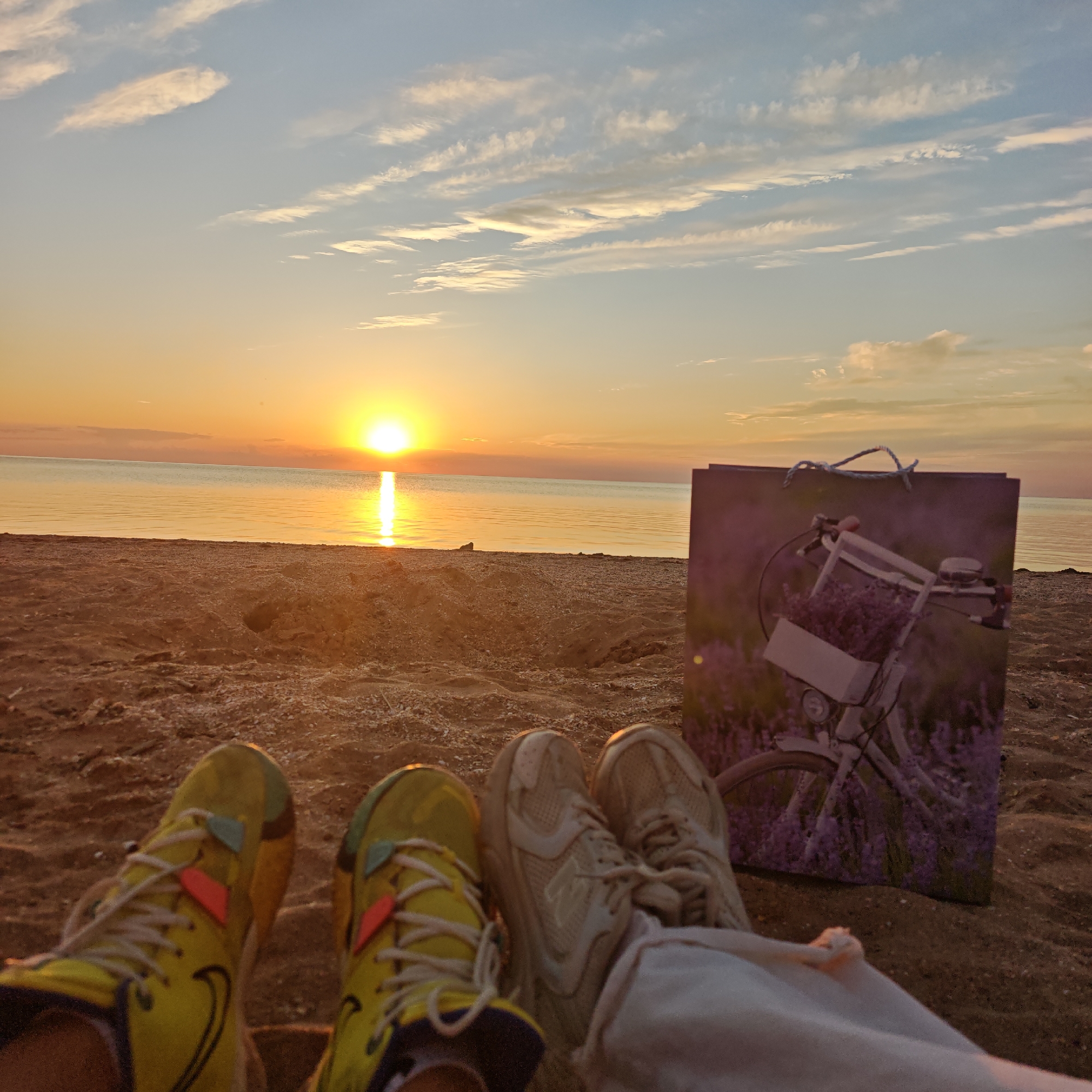 The image depicts a low-angle view of a coastal sunset or sunrise in Останинское сельское поселение, Ukraine, from the perspective of two individuals seated on a sandy beach. The sun, a bright orange disc, is positioned low on the horizon, slightly left of the image's vertical center, casting a vivid, straight reflection across the calm, golden-hued water surface. The sky transitions from a pale blue at the top to warm orange and yellow tones near the horizon, with scattered, elongated white and pale orange clouds.
In the mid-ground, a sandy beach extends from the water's edge. The sand is light brown with visible pebbles and shell fragments, and shows signs of disturbance, including small depressions.
In the foreground, the lower legs and feet of two individuals are visible. The person on the left wears bright yellow athletic shoes with white laces, subtle dark accents, and small triangular patches (one light blue-green, one orange-red) on the lacing. The skin of their lower leg is visible. The person on the right wears light beige/off-white athletic shoes with matching laces and light-colored (likely white or cream) pants that partially obscure their ankles. Both individuals appear to be facing the water and the setting/rising sun.
To the right of the individuals' feet, a purple-toned shopping or gift bag stands upright in the sand. The bag features an image of a vintage-style bicycle with a basket full of lavender flowers. A blue braided rope handle is visible at the top of the bag.
