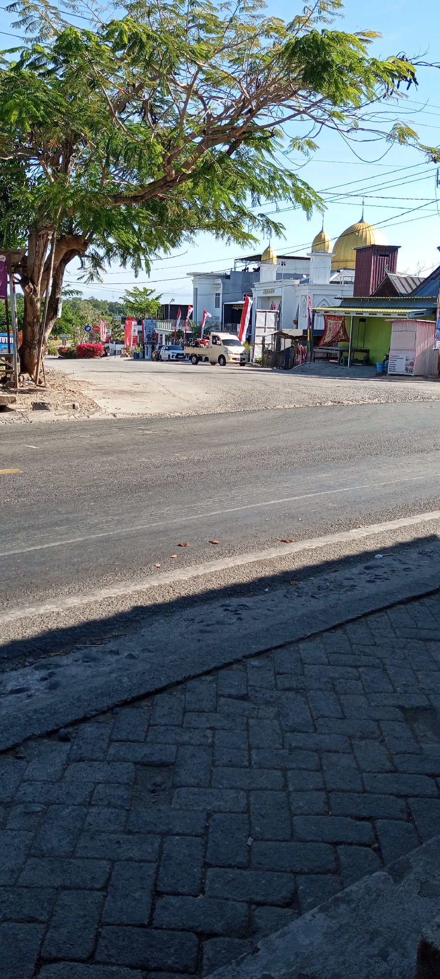 The image depicts a street scene in the city of Pakkanna, Indonesia. In the foreground, a paved road with a shadowed section on the right is visible. The road is bordered by a stone-paved sidewalk. Beyond the road, a large tree with green foliage stands to the left, casting a shadow on the paved area. In the mid-ground, several buildings are present, including a white mosque with golden domes, adorned with Indonesian flags. A light-colored truck is parked near the mosque. Various stalls and smaller structures line the street, some displaying banners and merchandise. The background shows a clear blue sky. The scene appears to be a typical street view in a town, with elements of commerce and religious architecture.