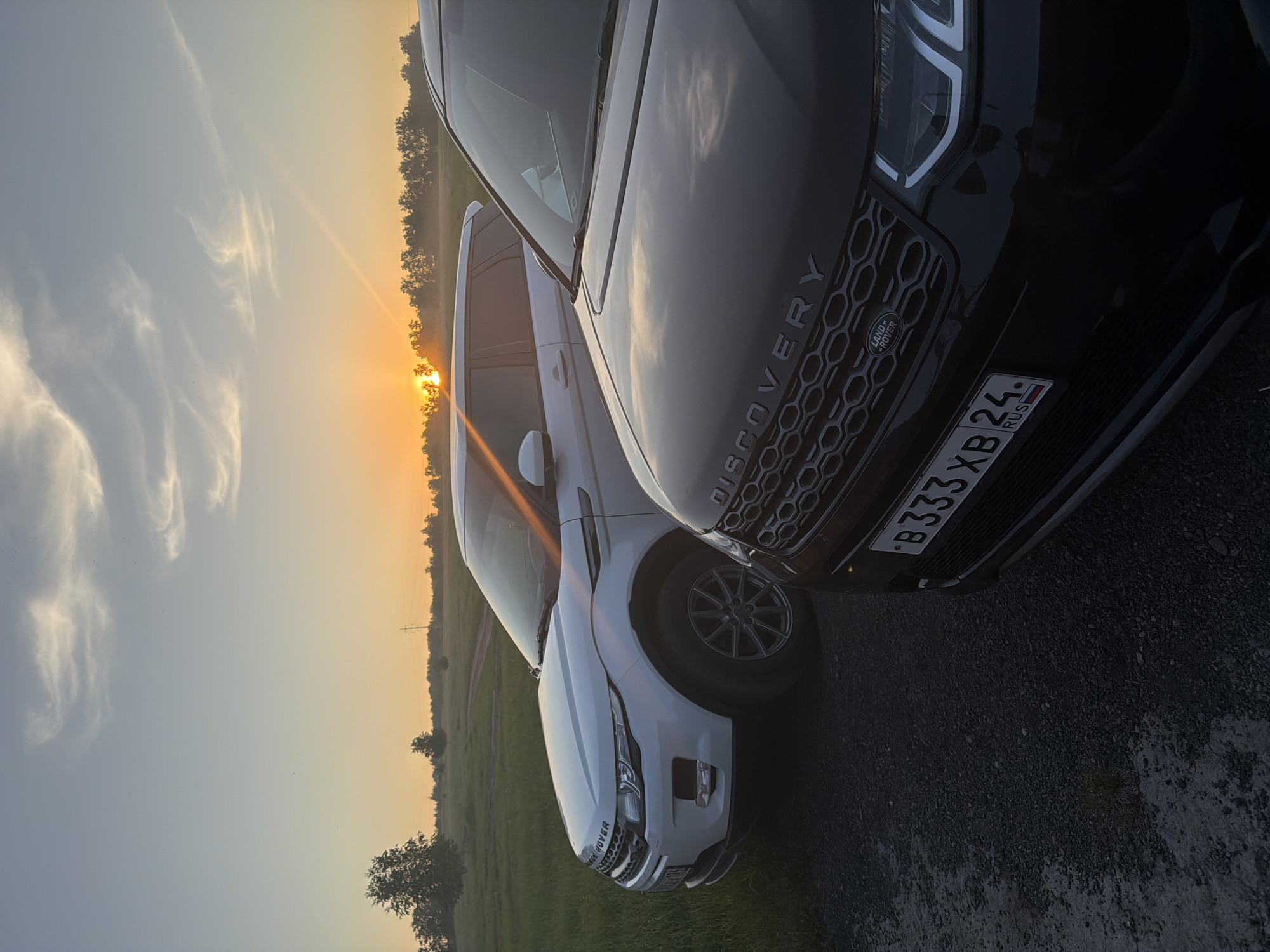 The image captures two Land Rover sport utility vehicles, one black and one white, parked on a gravel path during a sunset in a rural or semi-rural environment, specifically within Krasnoyarsk, Russia.
In the immediate foreground on the right, a black Land Rover Discovery Sport is prominently featured, displaying its front right quarter, hood, and grille. The word "DISCOVERY" is embossed on the hood, and the Land Rover emblem is centrally located on the grille. A Russian license plate, bearing the identifier "B333XB 24 RUS", is affixed to the front bumper; the "24 RUS" regional code indicates Krasnoyarsk Krai.
Situated behind and to the left of the black SUV, a white Range Rover Evoque (first generation) is partially visible. Its front left quarter, including the headlight assembly and a portion of the grille with "RANGE ROVER" branding, is discernible.
The vehicles rest on a dark gravel path that extends into a lighter dirt track, traversing a large expanse of green grassland. The horizon features a dense treeline, with individual trees scattered across the fields. A power line tower is visible in the far distance on the left, rising above the treeline.
The sky exhibits a warm, golden-orange hue near the horizon, typical of a sunset, with the sun partially visible and emitting a pronounced lens flare that traverses the upper section of the vehicles. The sky transitions to a lighter blue-grey overhead, containing several elongated, wispy cirrus clouds. The scene is backlit by the setting sun, casting warm light and creating contrast. No human figures are present.