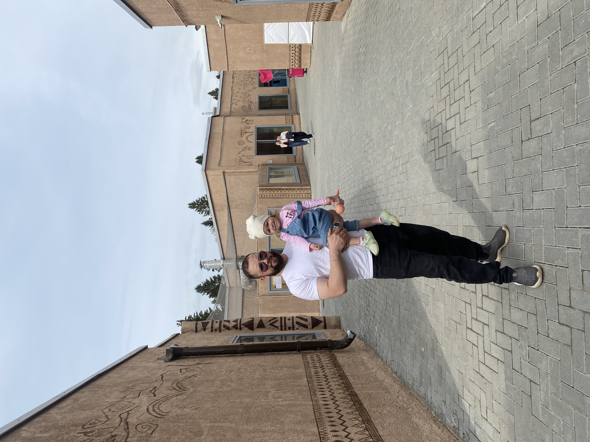 A man is holding a young girl up in the air in a courtyard. The man is wearing a white t-shirt, black pants, and sunglasses. The girl is wearing a white hat, a pink shirt, and a denim dress. The courtyard is paved with gray brick pavers. In the background, there are buildings with unique, tribal-like art designs. The location appears to be a courtyard or alleyway in the city of Perm, Russia, judging from the architectural style and art on the buildings. There are two additional people in the background, at a distance, near one of the buildings.