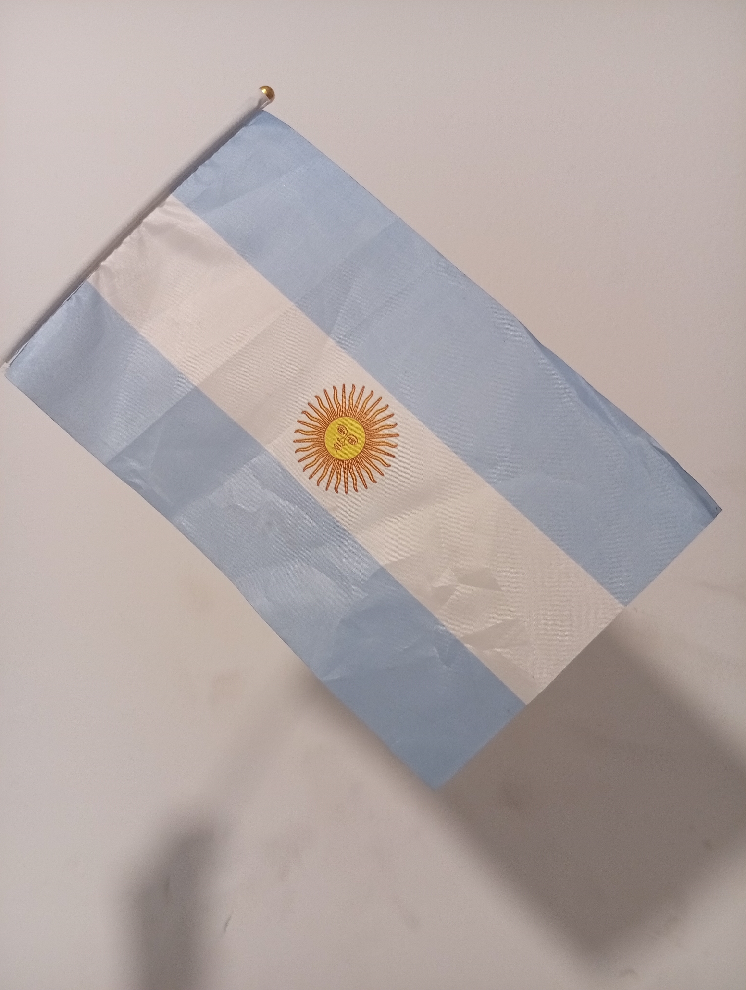 The media file showcases an Argentinian flag displayed against a plain, light-colored background. The flag features three horizontal bands: a light sky blue at the top and bottom, separated by a white band in the middle. Centered on the white band is the "Sun of May," a golden sun with a human face and 32 alternating straight and wavy rays, outlined in a reddish-brown color.
The flag appears to be made of a lightweight fabric, exhibiting soft creases and folds, suggesting it is either draped, gently held, or resting on a surface. A small, golden, spherical finial is visible at the top-left corner, indicating the flag is likely attached to a miniature flagpole. Soft shadows are cast by the flag onto the background, particularly noticeable towards the bottom right, implying illumination from an upper-left source. No people or activities are visible, and there is no discernible text on the flag or in the background. The scene is set in Buenos Aires, Argentina.