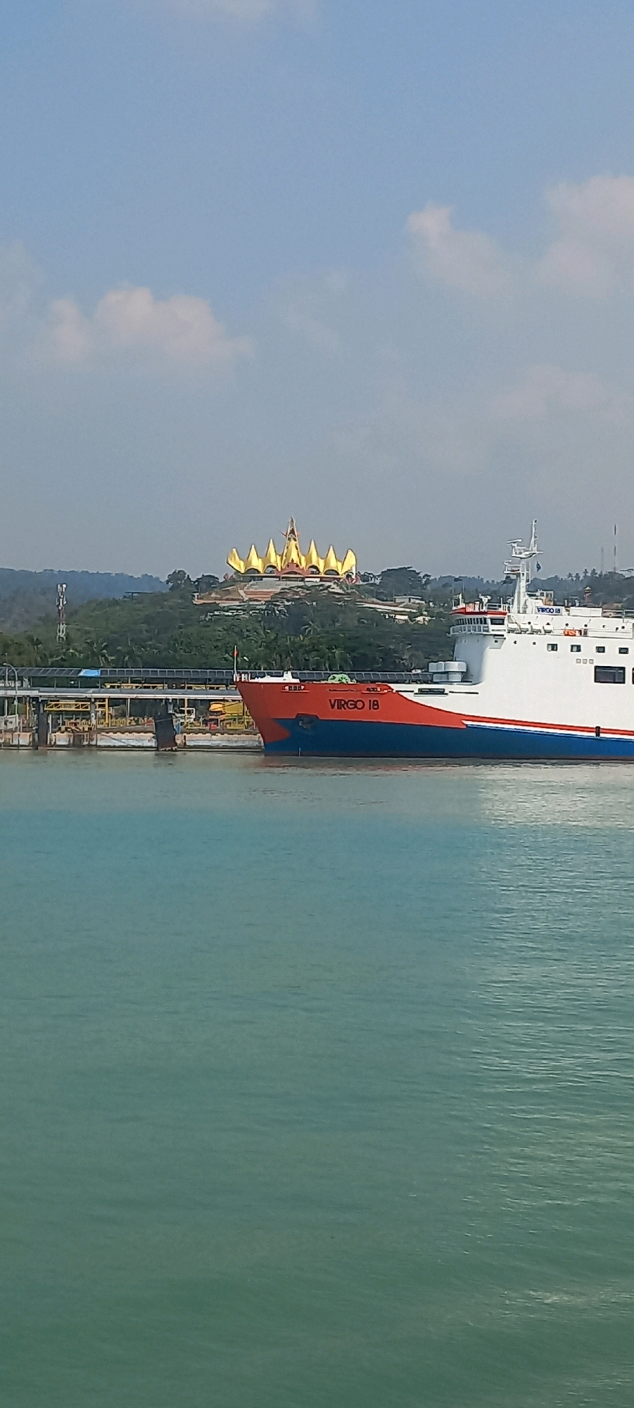The image shows a ferry named "Virgo 18" docked in the waters of Bakauheni, Indonesia. The ferry, colored red and blue, is positioned in the foreground. In the background, on a hill overlooking the water, stands a distinctive golden structure with multiple pointed spires. The surrounding landscape consists of lush green vegetation and some buildings. The sky is a clear blue with a few scattered white clouds. The water is a tranquil turquoise color, reflecting the bright sunlight.