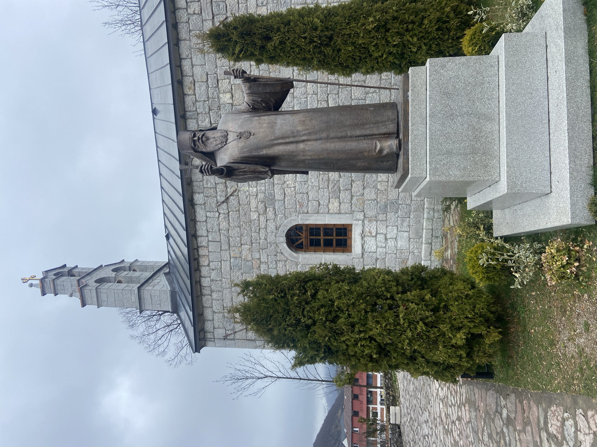 The image shows a bronze statue of a robed figure, possibly a religious figure, standing in front of a stone building. The statue is positioned on a tiered granite base. Behind the statue is a stone building with a tower and an arched window. Two tall, green, coniferous trees flank the statue. A stone-paved path leads away from the statue, and a building with red and white elements is visible in the background. The sky is overcast. This location is in Kolašin, Montenegro.