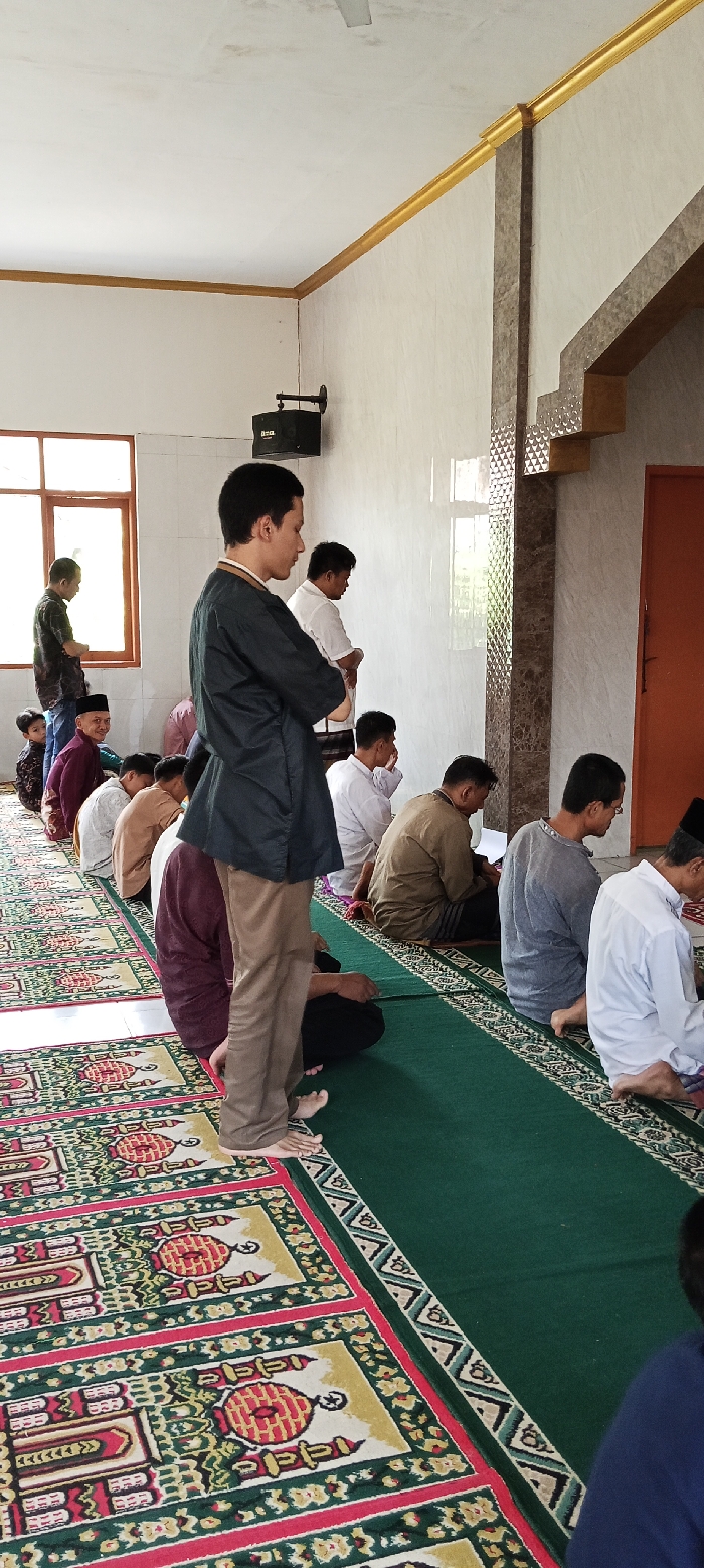 The image shows a room with a green carpet covering the floor. There are several men sitting on the carpet, with one man standing in front of them. The men are all wearing long-sleeved shirts and pants. The man standing in front of the group is wearing a black long-sleeved shirt and brown pants. The carpet is covered with prayer rugs, with intricate designs and patterns of red, yellow, and green. The walls of the room are white and decorated with a brown border. The room is well-lit and the walls are clean.
The location of the image is a room in a mosque in Kecamatan Kutawaringin, Indonesia, based on the provided coordinates (-6.973, 107.521).