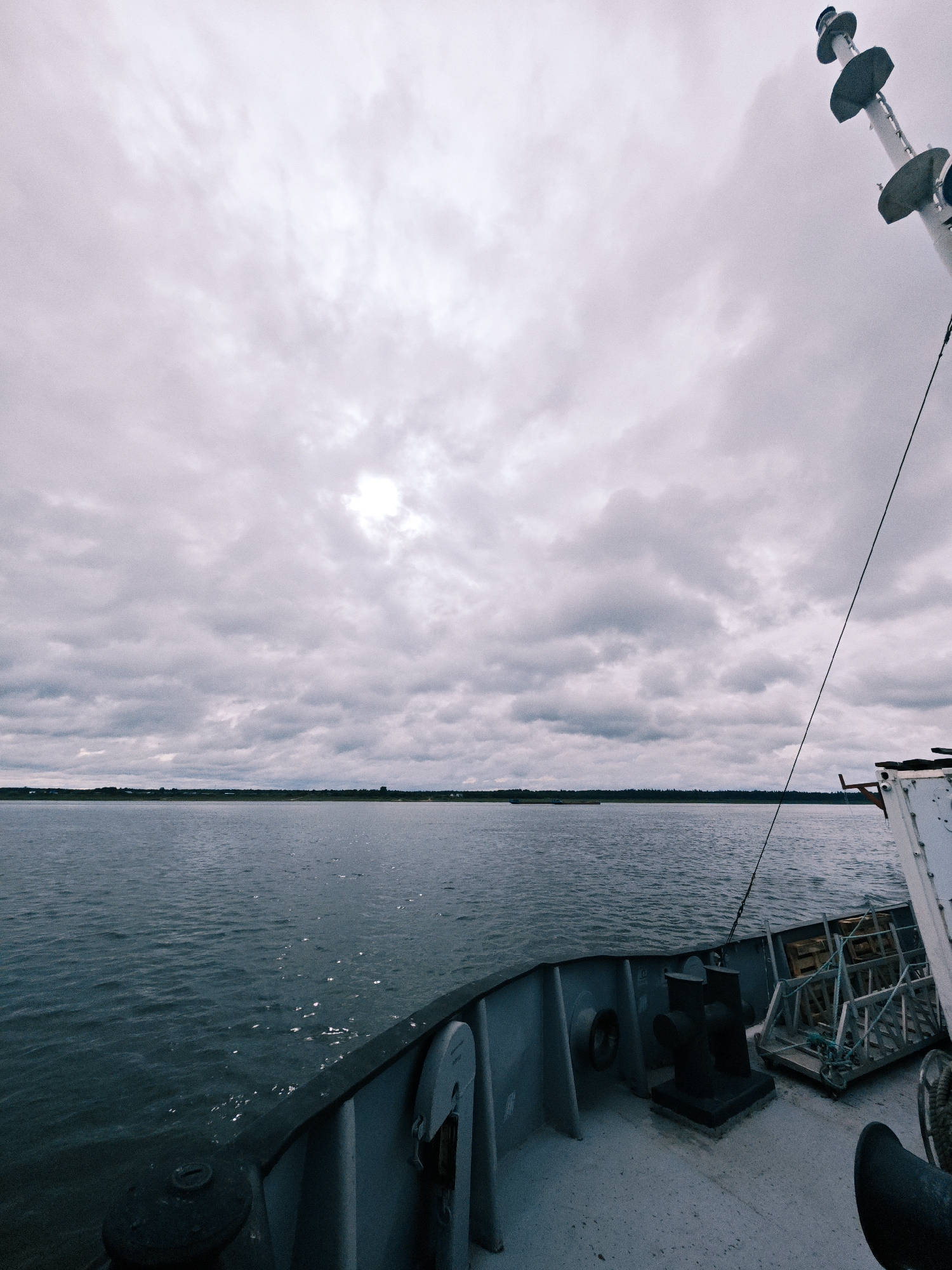 This is a low-angle, full shot of the bow of a vessel on a body of water under a cloudy sky. The foreground features the deck of the ship, with a dark, bollard-like structure on the left and a lighter, rectangular structure with a winch on the right. The midground is dominated by a wide expanse of rippling, dark blue water. In the distance, a flat horizon of dark green trees and some low structures, likely buildings, stretches across the frame. The sky is filled with heavy, gray clouds, with a bright, diffused patch of sunlight near the center, suggesting an overcast day. A tall, white mast with multiple dark, circular platforms is visible on the far right of the image, extending upwards into the sky. A thin black cable runs diagonally from the mast down towards the left side of the frame. The overall atmosphere is muted and somber due to the cloudy weather. The location in Zotino, Russia, indicates a likely setting on a river or lake.