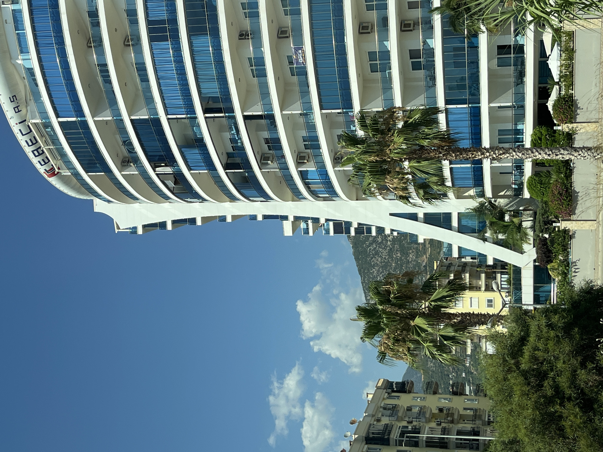 The image shows a modern, curved white building with multiple balconies featuring blue glass railings and blue curtains. The building has "CEBECI A.S." written on its top in red letters. The building is located in Alanya, Turkey, as indicated by the presence of palm trees and the architectural style common in coastal Turkish cities. To the left of the main building, there is a yellow apartment building with balconies. In the background, a tree-covered mountain is visible under a clear blue sky with scattered white clouds. The foreground shows lush green trees and a stone wall. White umbrellas are visible on the ground level of the main building.