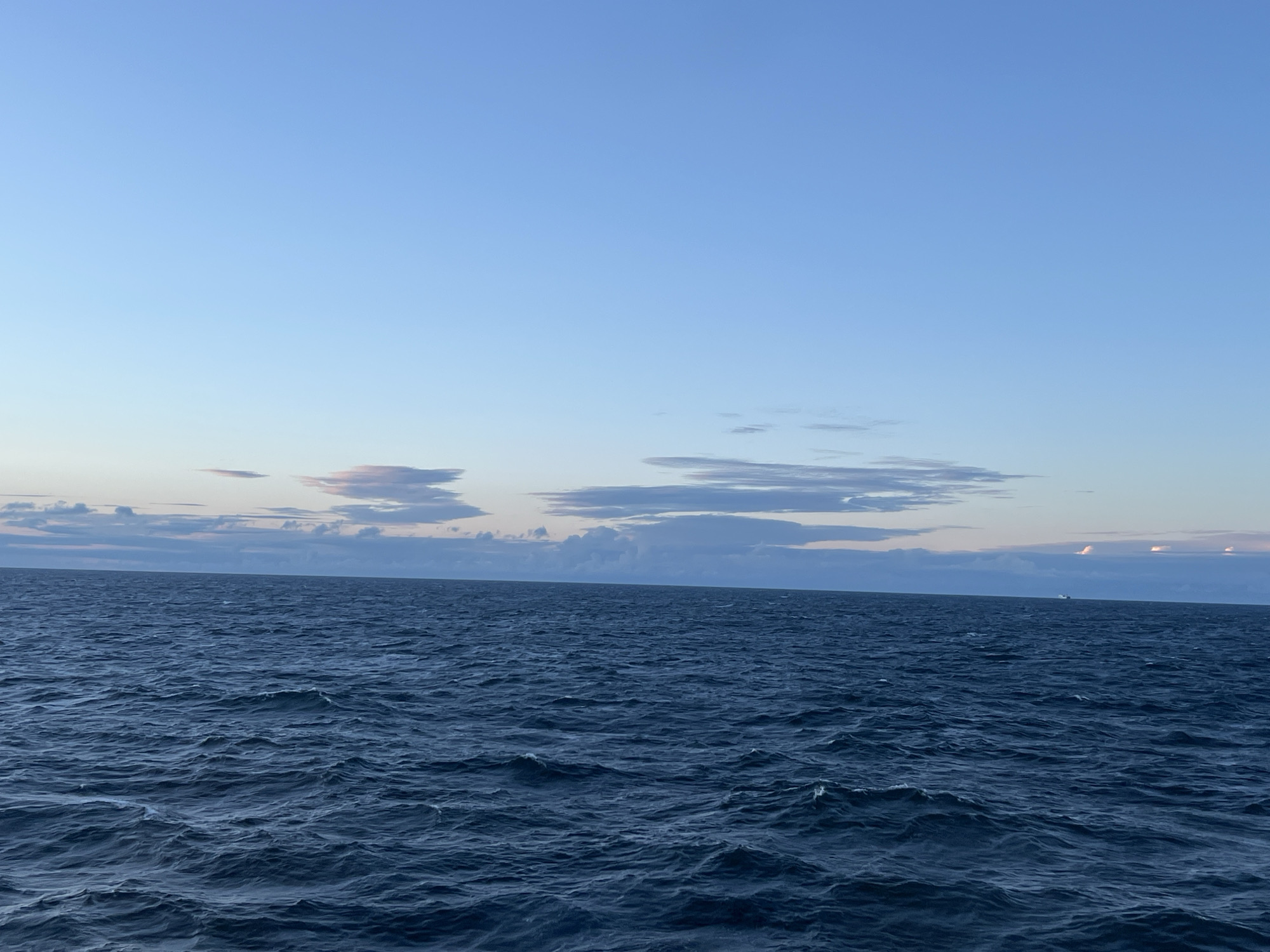 An image depicts a vast expanse of deep blue ocean under a pale blue sky. A faint horizon line separates the water from the sky, where scattered wispy clouds are visible, tinged with soft pinks and purples, suggesting either dawn or dusk. The ocean surface is active with moderate waves, creating a textured and dynamic foreground. The water appears dark and deep, reflecting the cool tones of the sky. There are no people or specific landmarks visible that would indicate a location within the city of Unknown, Poland. In the distance, a small, faint object, possibly a ship or buoy, is visible on the horizon.