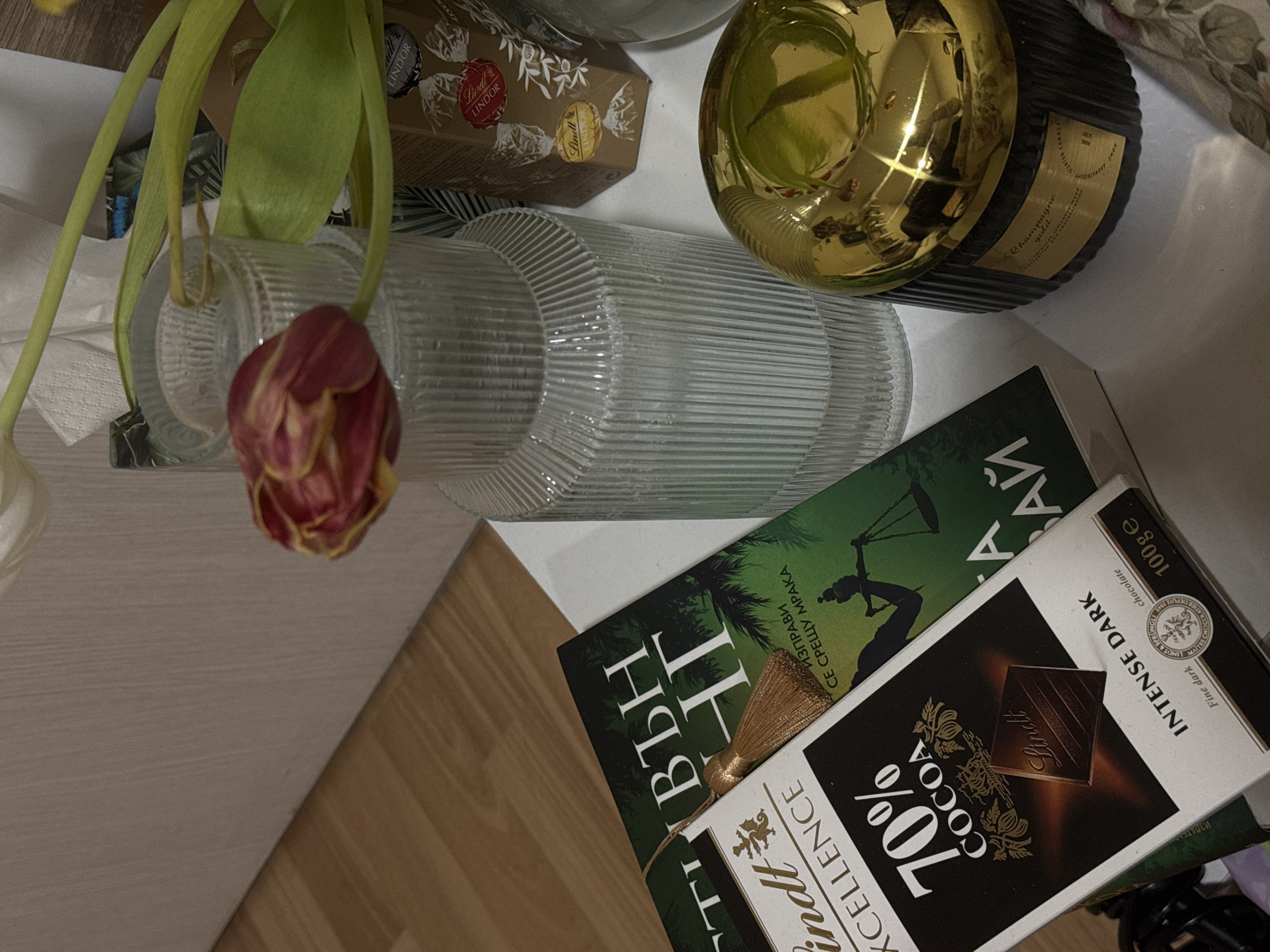 An indoor, static scene displays various items arranged on a white, flat surface positioned against a light brown, vertically textured wall. A wooden floor or adjacent surface is visible to the lower left.
Subjects and objects present include:
* One clear, ribbed glass vase holding two tulips; one white and one red/yellow striped, the latter appearing partially wilted.
* One dark green hardcover book with Cyrillic text, reading "СПРАВЕДЛИВЪН ЗАЙ" (Justice for the Bear) and "ИЗПРАВИ СЕ СРЕЩУ МРАКА" (Face the Darkness) attributed to Michael Connelly. A gold-tasseled bookmark is inserted into the book.
* One 100g Lindt Excellence 70% Cocoa Intense Dark chocolate bar, partially covering the book.
* One cylindrical black candle jar with vertical ridges and a shiny gold, ribbed lid. A label on the jar reads "Champagne gold scented candle."
* One small, hexagonal cardboard box of Lindt Lindor chocolates, brown with red and gold branding, located behind the vase.
* A crumpled white paper napkin or tissue is visible behind the vase and book.
* A fragment of floral-patterned fabric is visible on the far right side of the white surface.
No people are present. There are no active interactions; the scene is a still life arrangement.
The presence of a book with Bulgarian text (Cyrillic script, Bulgarian title and author) indicates the scene is located in a generic indoor setting, likely a private residence or other interior space, within the city of Sofia, Bulgaria.