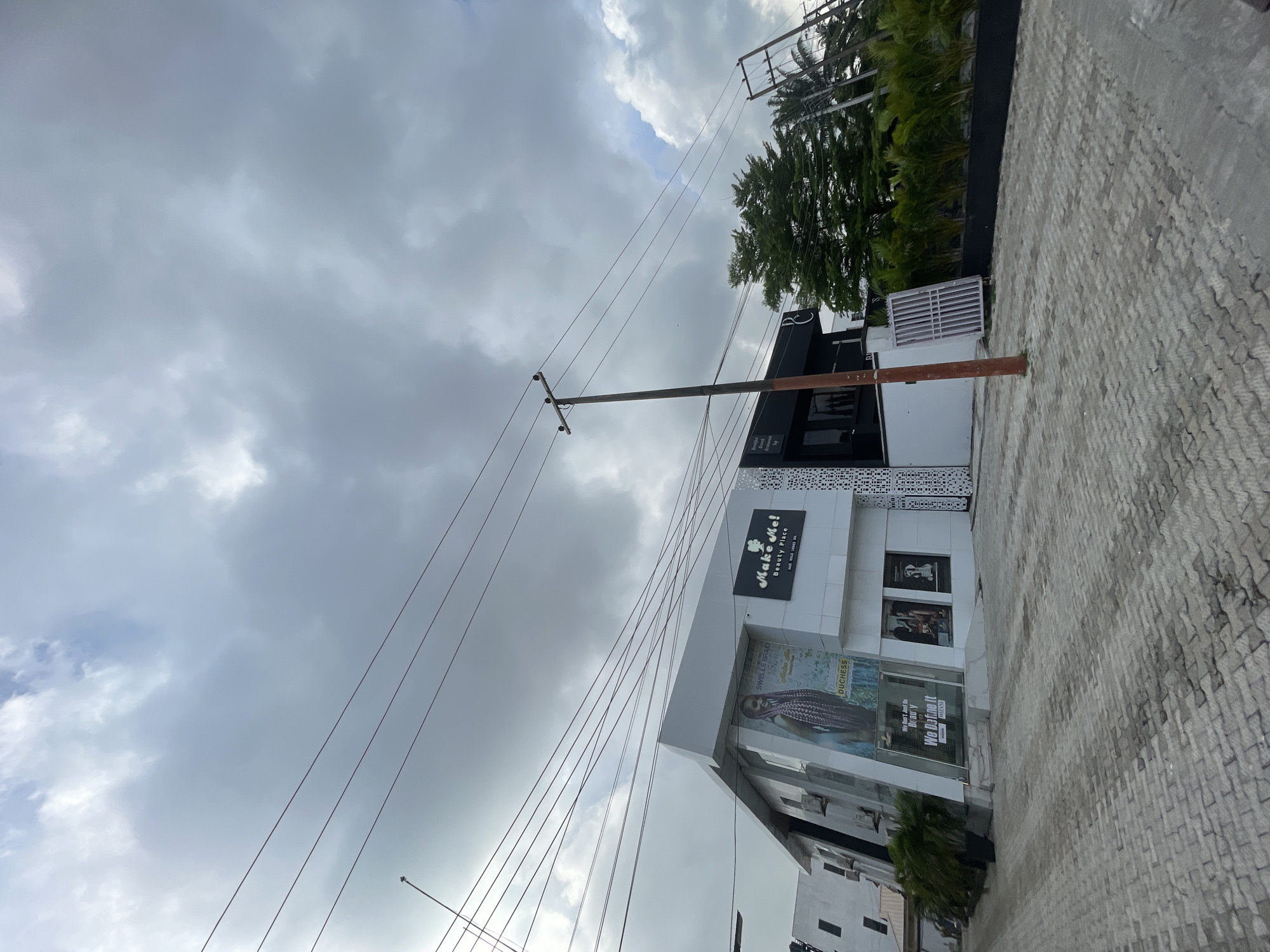 The image shows a building with the sign "Make Me! Beauty Place" in Moba, Nigeria. There is an advertisement for "SWELLS BRAD Duchess" and another sign with the text "We Define it MIZANI" in the window. The sky is cloudy and there are power lines visible in the foreground. The street in front of the building is paved with stone blocks. Some trees and greenery are visible next to the building.