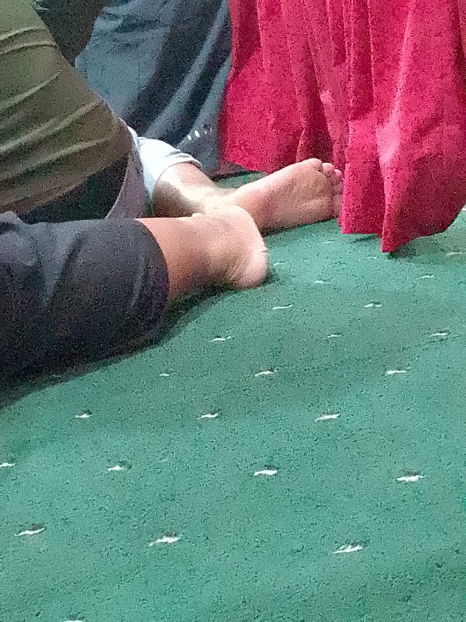 In Kertajaya, Indonesia, an image captured from a low angle shows a person's bare feet resting on a green carpet. The carpet has a subtle pattern of small, elongated white dashes. To the left, a person's legs are visible, clad in dark pants and what appears to be an olive-green garment. White fabric, possibly a sock or bandage, is wrapped around one ankle. A large piece of bright red fabric, possibly a curtain or draped cloth, hangs to the right, obscuring the background and the rest of the scene. The lighting suggests an indoor setting.
