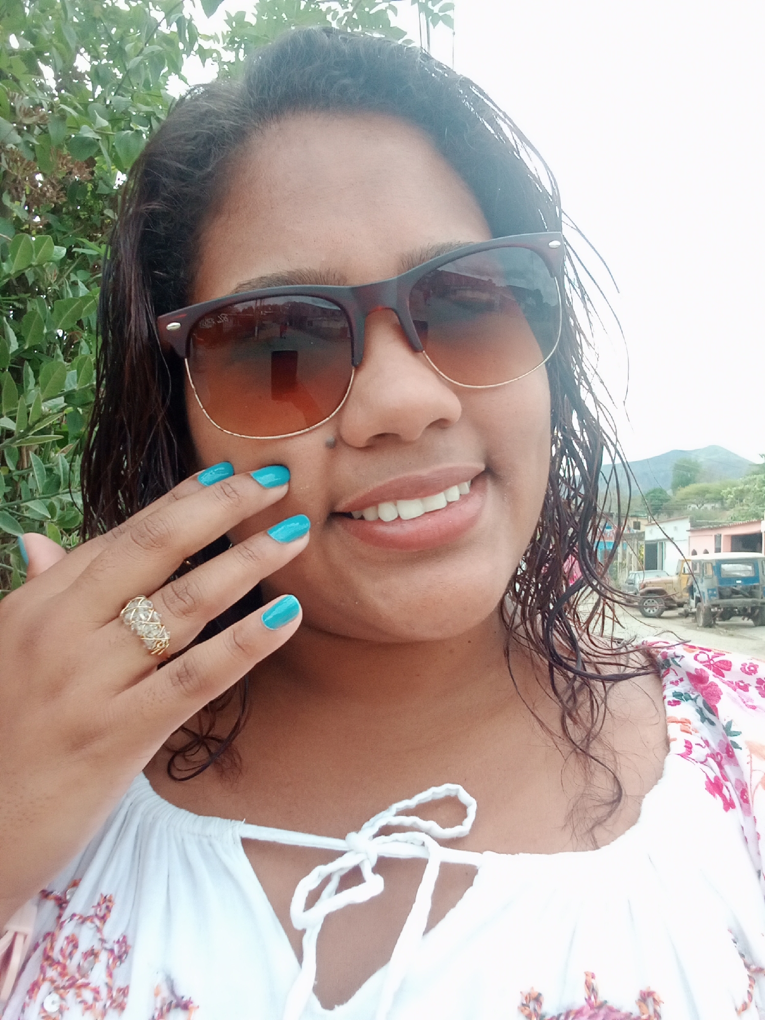 A woman is shown in a selfie-style close-up. She wears brown-framed sunglasses and has wet, dark hair. Her fingernails are painted bright blue. She has a gold ring on her ring finger and is wearing a white blouse with floral patterns on the sleeves. The background includes greenery on the left side and houses, vehicles, and a mountain in the distance, indicating an urban setting within the Parroquia Ocumare, Venezuela.