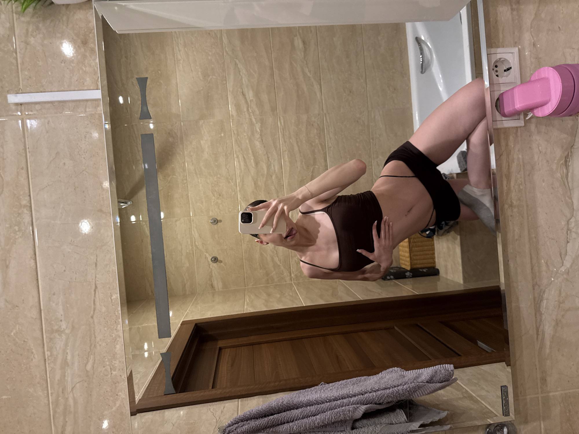 A woman takes a mirror selfie in a bathroom. She is wearing a dark brown tank top and dark shorts, with one hand on her stomach and the other holding a phone. Her tongue is sticking out and her eyes are closed. She is posing with her legs bent and her body tilted. The bathroom features beige tiled walls, a wooden door, a towel rack with a gray towel, and a white bathtub. A pink electrical adapter is plugged into an outlet near the bottom right of the frame. The image appears to be taken in an indoor residential bathroom, possibly in the city of Khimki, Russia.