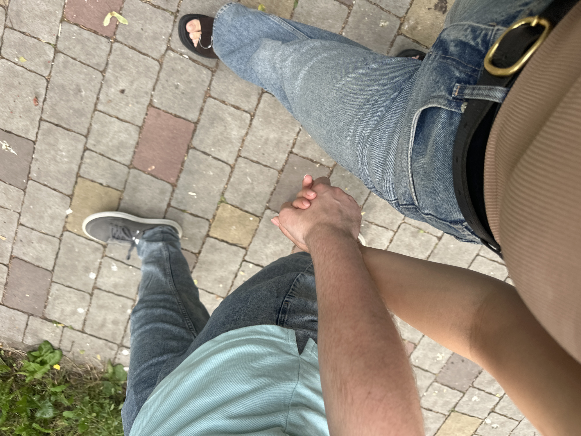 The image shows a close-up, high-angle view of two people holding hands while walking on a paved walkway in Ternopil, Ukraine. The walkway is constructed of rectangular interlocking pavers in shades of gray, red, and yellow. To the left of the walkway, there is a patch of green grass with some dry leaves and debris.
The person on the left is wearing a light blue polo shirt and dark wash jeans. Their right arm is extended, and their hand is clasped with the other person. They are wearing gray sneakers with white laces. The person on the right is wearing blue jeans and a tan top that is partially visible. They are wearing black sandals with a single strap across the toes and a buckle. The hands are clasped loosely, suggesting a casual stroll. The overall impression is that of a couple enjoying a walk outdoors. The paved walkway and surrounding greenery are typical of an urban park or pedestrian area in Ternopil.