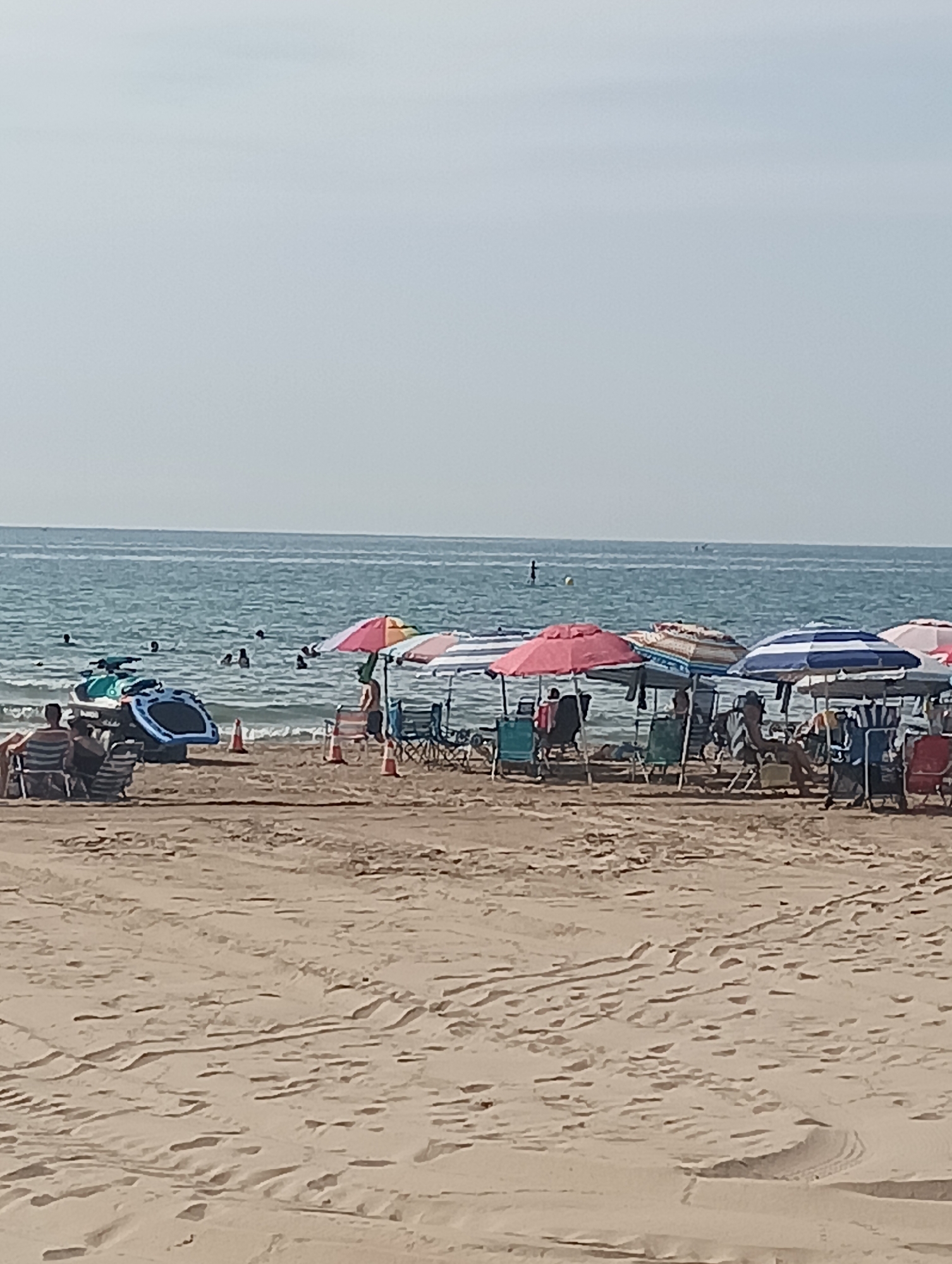 The image depicts a beach scene in La Playa, Spain. Several people are visible in the water, swimming or enjoying the ocean. On the sandy shore, a group of beachgoers are seated in chairs under various colorful umbrellas, including red, pink, white, and blue striped umbrellas. Some are relaxing, while others appear to be conversing. In the foreground, the sand is marked with tire tracks, indicating vehicular or equipment movement. There are also some inflatable water toys and traffic cones on the beach. The sea extends to the horizon under a pale, overcast sky.