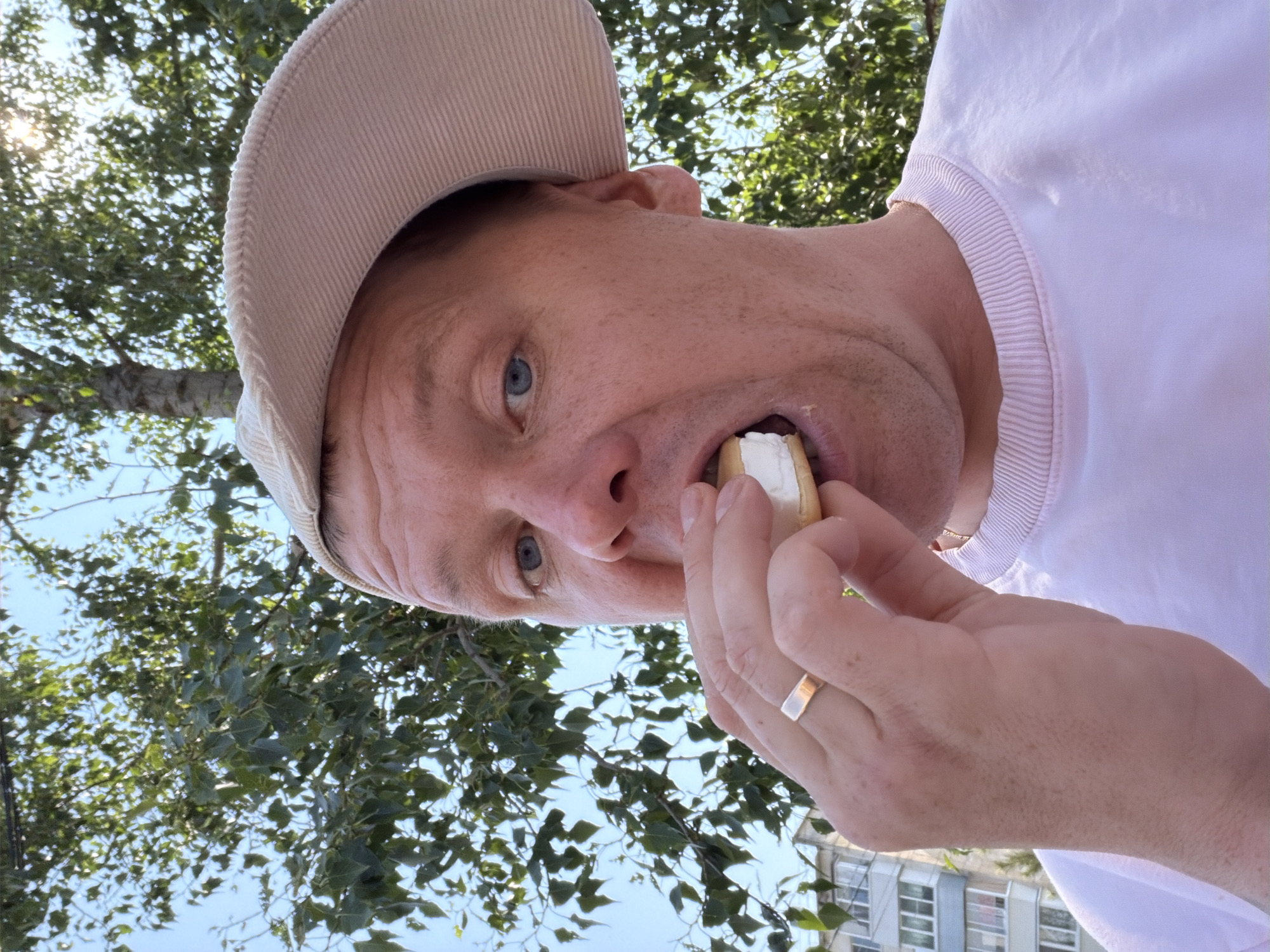A Caucasian man with light skin, blue eyes, and sparse facial hair is wearing a light-colored baseball cap and a light pink crewneck sweatshirt. He is holding a sandwich cookie with white cream filling up to his mouth and taking a bite. He has a gold ring on his left ring finger. The man is outdoors, with green foliage and a bright blue sky visible in the background, indicating it is daytime. Sunlight is filtering through the trees. To the left of the man, a building with multiple stories and balconies is visible, consistent with a residential building in an urban or suburban environment. The building is light-colored with some blue accents. The setting appears to be the city of Chaikovsky, Russia, given the characteristic architecture and foliage.