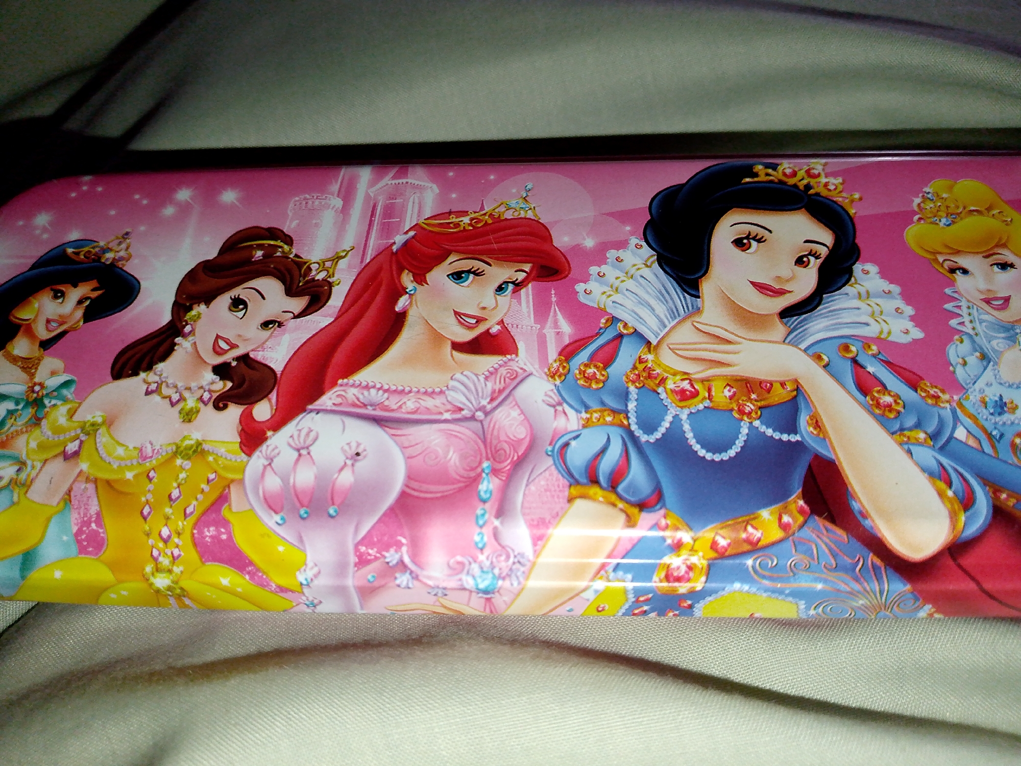 The image shows a close-up of a colorful tin pencil case featuring an illustration of five Disney princesses. From left to right, the princesses are likely Jasmine, Belle, Ariel, Snow White, and Aurora, all depicted in their signature gowns and with crowns. They are standing in front of a pink, sparkly background with illustrations of castles. The tin appears to be resting on a textured, light-colored surface, possibly fabric. The lighting suggests an indoor setting.