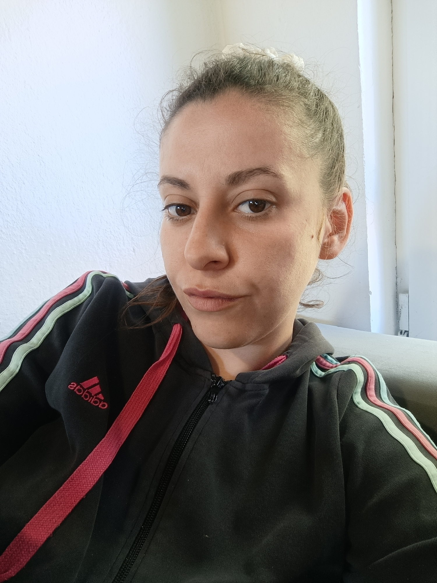 A woman is seated, wearing a zipped-up Adidas track jacket with pink drawstrings and pink and mint green stripes down the sleeves. Her hair is pulled back. She has brown eyes and fair skin. The background is a plain white wall, possibly in a residential setting. There is a slight suggestion of a couch to the lower right. The image appears to be a self-portrait. The image provides no information to verify the location in Gorna Rositsa, Bulgaria.
