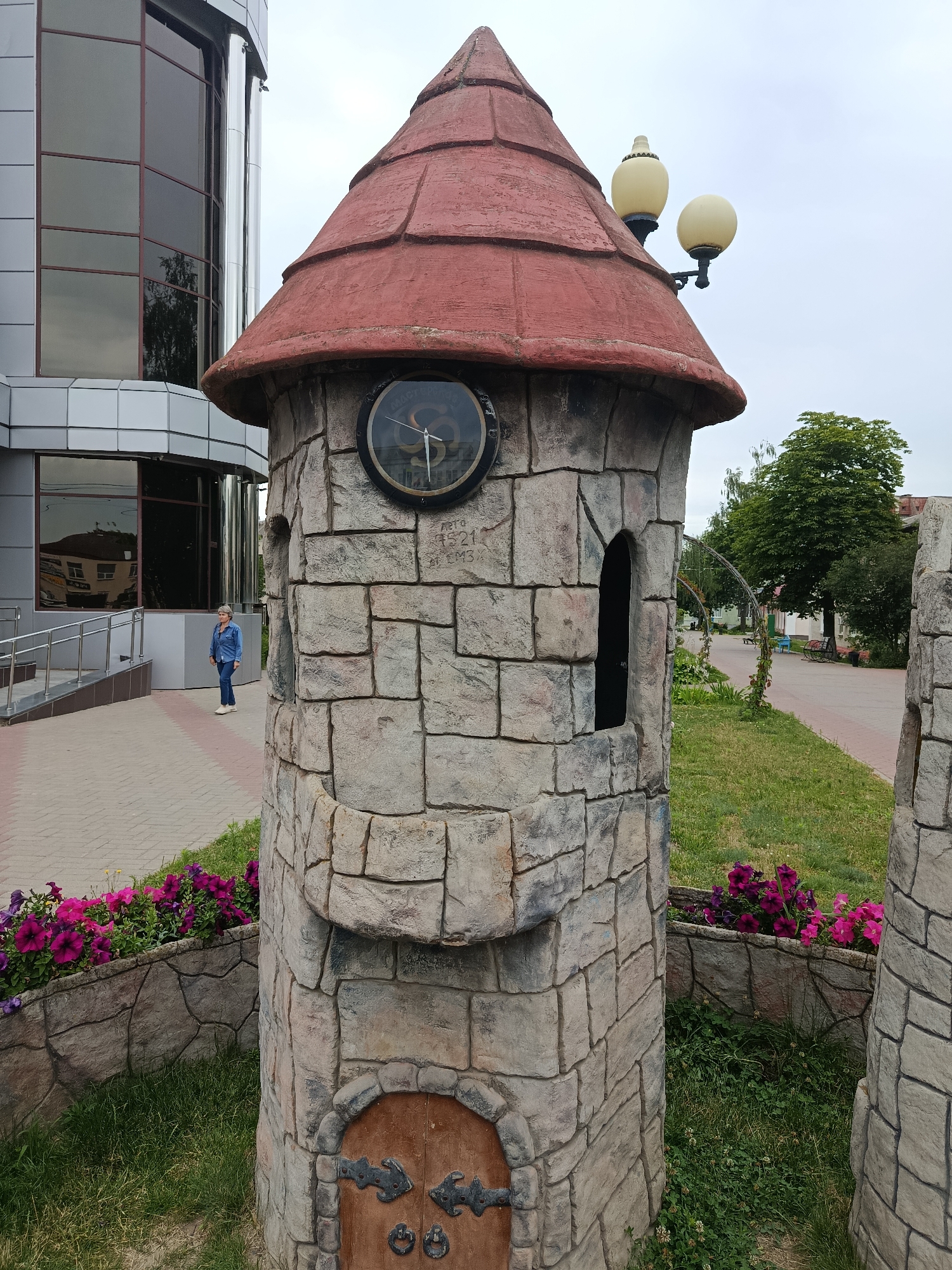 The image depicts an outdoor scene in the city of Zhlobin, Belarus. In the foreground, a decorative miniature castle tower made of stone-like material stands prominently. It features a red conical roof, a clock face with Roman numerals and hands, and small arched window openings. At the base of the tower is a small wooden door with decorative metal hinges. In front of the tower and extending to the left is a low stone wall adorned with vibrant pink and purple flowers.

To the left of the tower, a modern building with large glass windows is partially visible. A woman wearing a denim jacket and jeans stands on a paved walkway in front of this building, facing away from the camera. The walkway is bordered by a gray metal railing and steps leading up to the building.

The background shows a park-like setting with a paved pathway, green grass, trees, and some park benches. Two globe-shaped streetlights are mounted on a pole near the top of the castle tower. The sky is overcast. The scene appears to be a public space, possibly a park or a square in front of a commercial or administrative building.