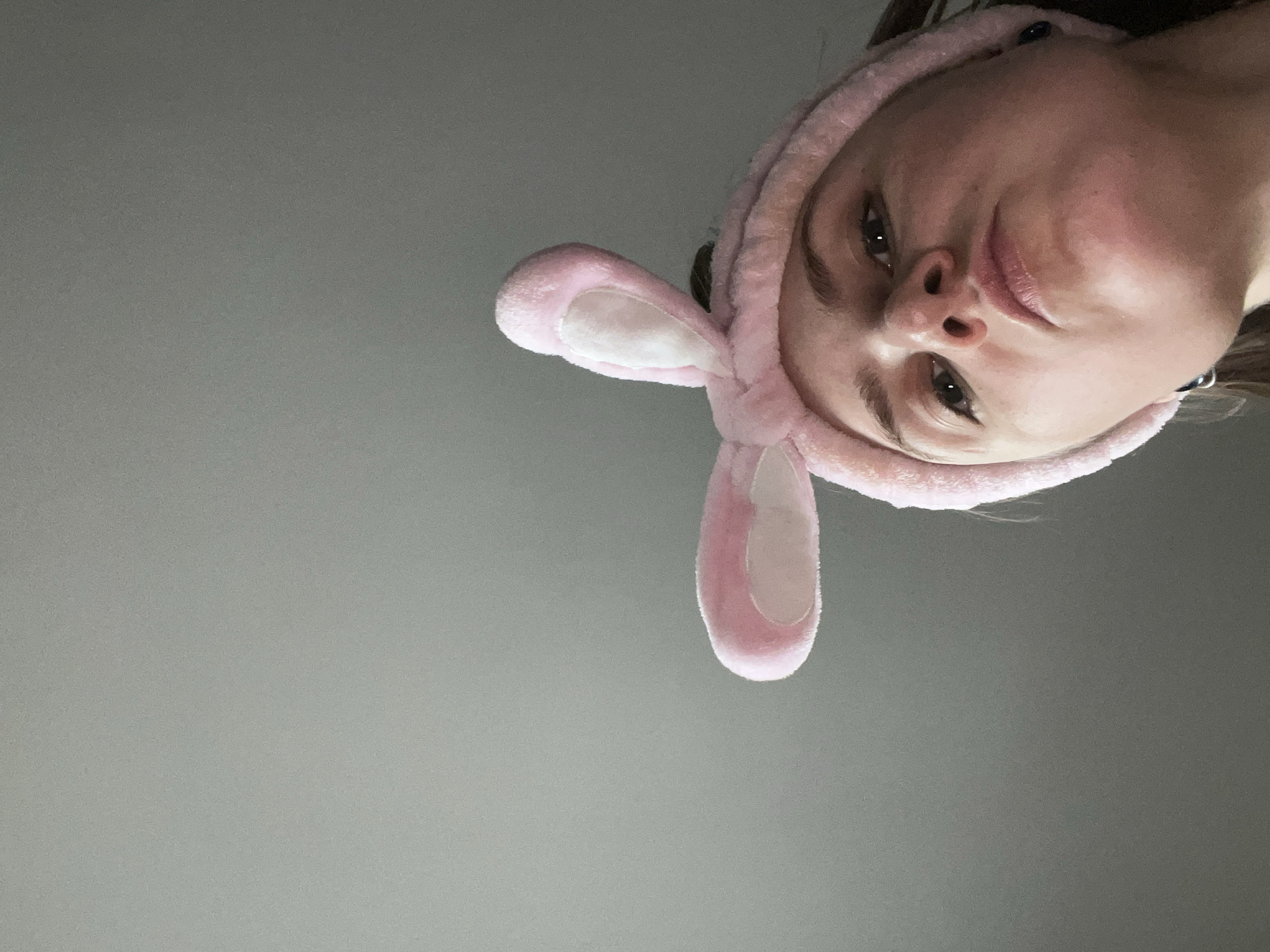 A low-angle shot depicts a person with fair skin wearing a pink bunny ear headband. The background is a plain, gray surface. The person's face is angled towards the camera, and their eyes are visible. The location is not discernible in the image.
