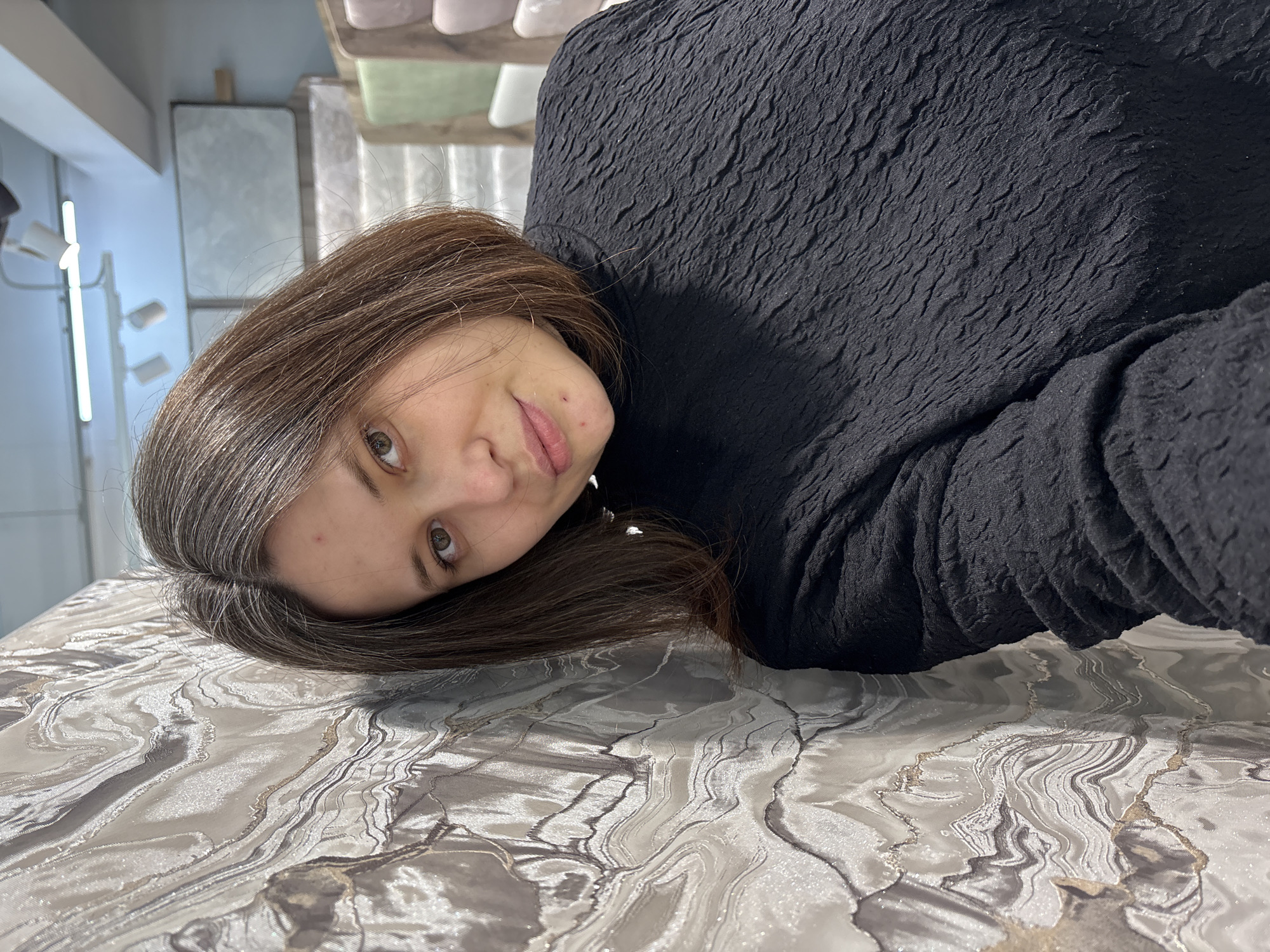 A woman with light brown hair is lying on a surface with a marble-like pattern. She has light skin and brown eyes. She is wearing a black, textured top. The location may be in Chaikovsky, Russia, although there are no visual cues to confirm this. The background includes a white wall, a light fixture, and blurry objects on a shelf. The photograph appears to be taken from a low angle, with the camera close to the woman's face.