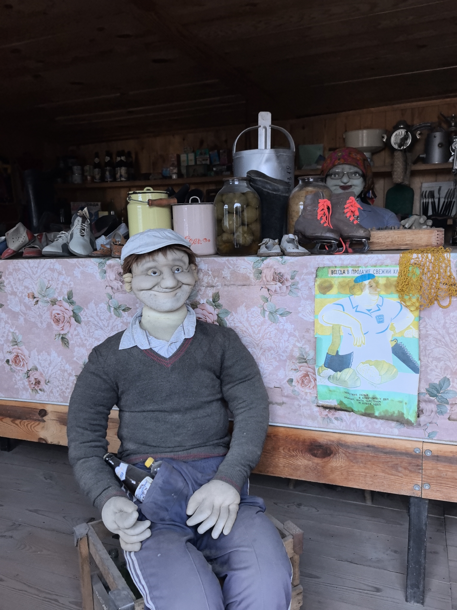 A staged display in Bol'shoi Mashlyak, Russia, shows a pale-skinned, male mannequin wearing a newsboy cap, a grey V-neck sweater, a blue button-down shirt, and blue sweatpants with white stripes. He sits on a wooden crate, holding a beer bottle in his left hand. The mannequin appears aged, with prominent wrinkles and a forced smile.
Behind the mannequin, a table covered with a pink floral-patterned cloth displays an eclectic array of items. These include shoes, ice skates with orange laces, jars of pickles, metal containers, and an elderly female mannequin wearing glasses and a floral headscarf. A vintage advertisement is pinned to the table, showing a baker and bread, with Cyrillic text. Various utensils and containers are visible on the shelves in the background. The setting appears to be a wooden shed or a booth of some kind.