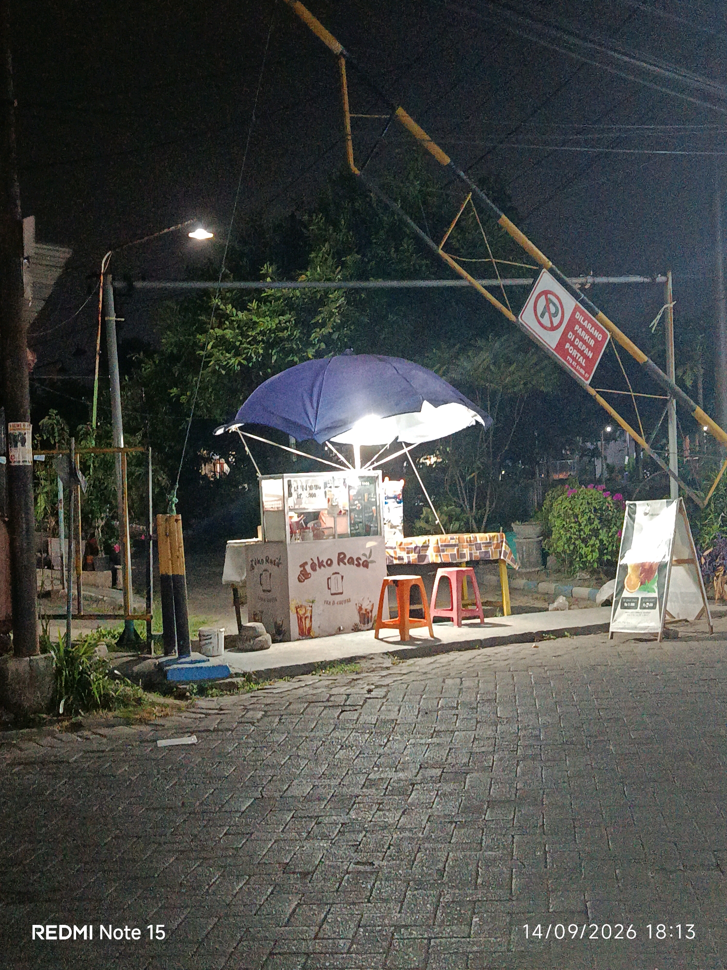 In Keboansikep, Indonesia, at night, a street food vendor stall is illuminated by a blue umbrella and lights. The stall, branded "Toko Rasa", displays images of drinks. Two stools, one orange and one pink, are placed in front of the stall. A white A-frame sign stands to the right of the stall, displaying drink advertisements. The stall is situated on a paved street, with a yellow and black metal barrier overhead. A red and white "No Parking" sign with text in Indonesian is attached to the barrier. Trees and bushes form the background. The bottom of the image contains the text "REDMI Note 15" and "14/09/2026 18:13".